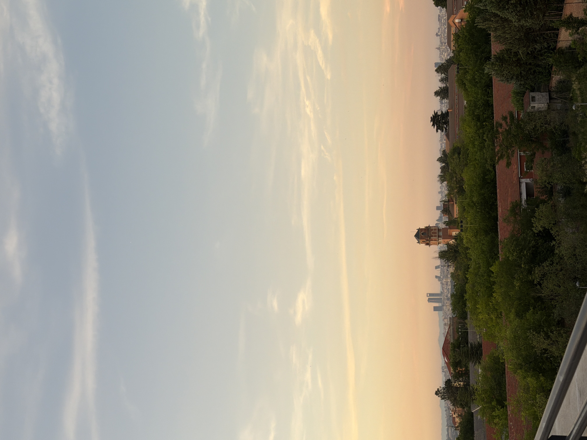The image captures a cityscape at dusk, with the sky transitioning from a pale blue at the top to warm hues of orange and yellow towards the horizon. A few wispy clouds are visible, catching the fading light. In the middle ground, a cluster of trees partially obscures a collection of buildings, characterized by red-tiled roofs. Prominent among these is a reddish-brown tower with a decorative top, appearing to be a historical structure. Beyond the immediate buildings and trees, a more distant skyline is visible, featuring several modern skyscrapers. The foreground is dominated by a railing, suggesting the viewpoint is from an elevated position, such as a balcony or rooftop. The overall scene conveys a sense of urban tranquility as day gives way to night.