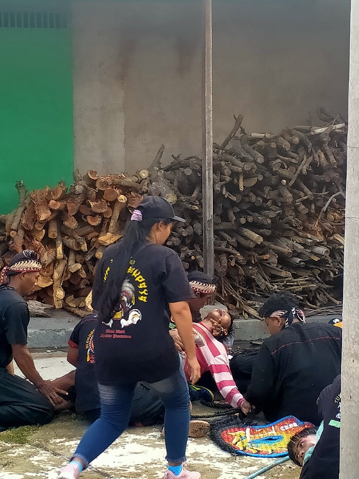 The image captures an outdoor or semi-outdoor scene in the city of Sepat, Indonesia. In the background, two large, separate piles of cut firewood and logs are stacked against a wall, which is partially painted green on the left and unpainted concrete on the right. A dark vertical column stands between the two wall sections and woodpiles.
In the mid-ground, a woman, viewed from behind, walks towards the right. She wears a black baseball cap, a black t-shirt with a colorful rooster graphic and text on the back, blue jeans, and pink athletic shoes with blue socks. Her long dark hair extends down her back.
To the right of the walking woman, a man in a pink and white striped long-sleeved shirt and a patterned headband is lying on the ground or heavily reclined. His head is tilted back, mouth open, and eyes partially closed, indicating an unresponsive or altered state. Surrounding him are at least two other individuals, both wearing dark clothing and patterned headbands, kneeling or sitting on the ground. One is to the left of the man in the striped shirt, and the other is to his right, with red stitching visible on their black top.
In the lower right corner, another individual is partially visible lying prone on the ground, wearing a dark shirt, with part of their face visible. Nearby, a brightly colored, multi-patterned mat or textile object lies on the ground. The ground surface appears to be a light-colored, possibly concrete, material with some darker, wet-looking areas. A black rope or cord is visible near the man in the striped shirt.