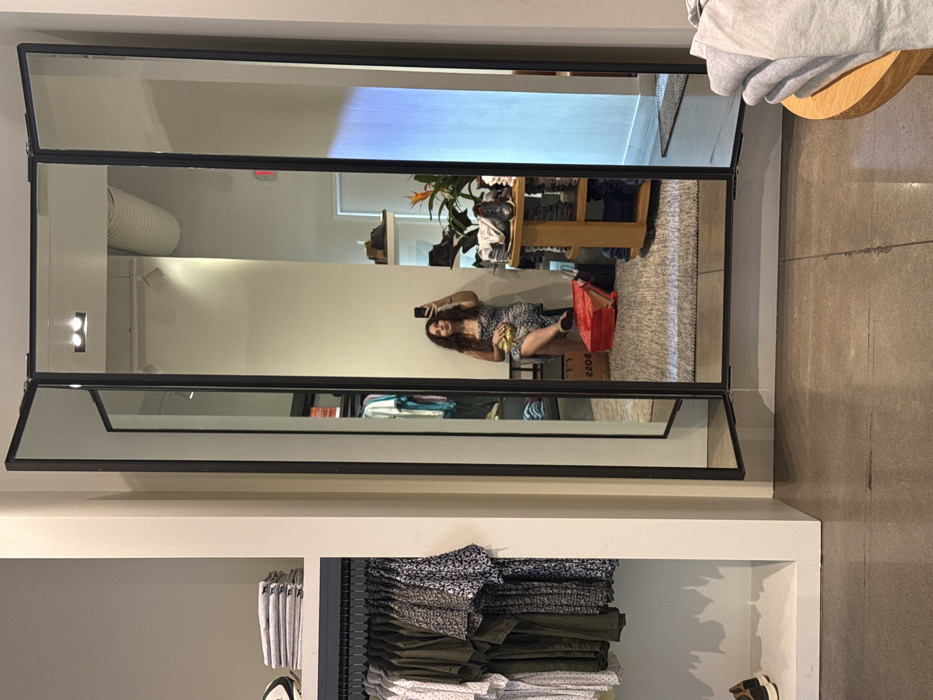 A hinged mirror in a retail store reflects a woman taking a selfie with her phone. She is sitting on a box in the reflection. The store has clothing racks and displays. The floor is concrete. A table with folded clothes sits to the right of the mirror.