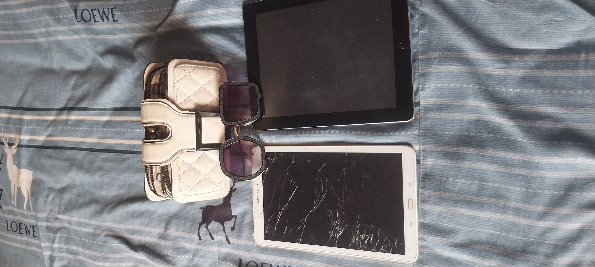 The image displays a white quilted handbag with zippers positioned above two tablet devices and a pair of sunglasses. One tablet, on the left, has a severely cracked screen. The other tablet, on the right, has a black screen. The sunglasses are placed in front of the tablets. The scene is set against a blue and white striped fabric, which appears to be a bedsheet, featuring a faint pattern of a deer. The image is taken from a top-down perspective. The location of Onitsha, Nigeria is not discernible from the visual information provided in the image.