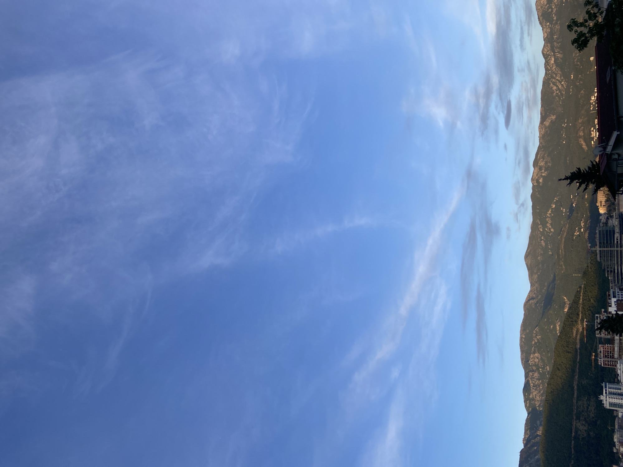The image shows a landscape with a clear, blue sky filled with wispy cirrus clouds on the left. On the right, a mountain range with green vegetation and rocky terrain forms a natural border. The bottom of the mountain features buildings, suggesting a city or urban area encroaching on the natural landscape. The location is in the city of Unknown, Unknown.