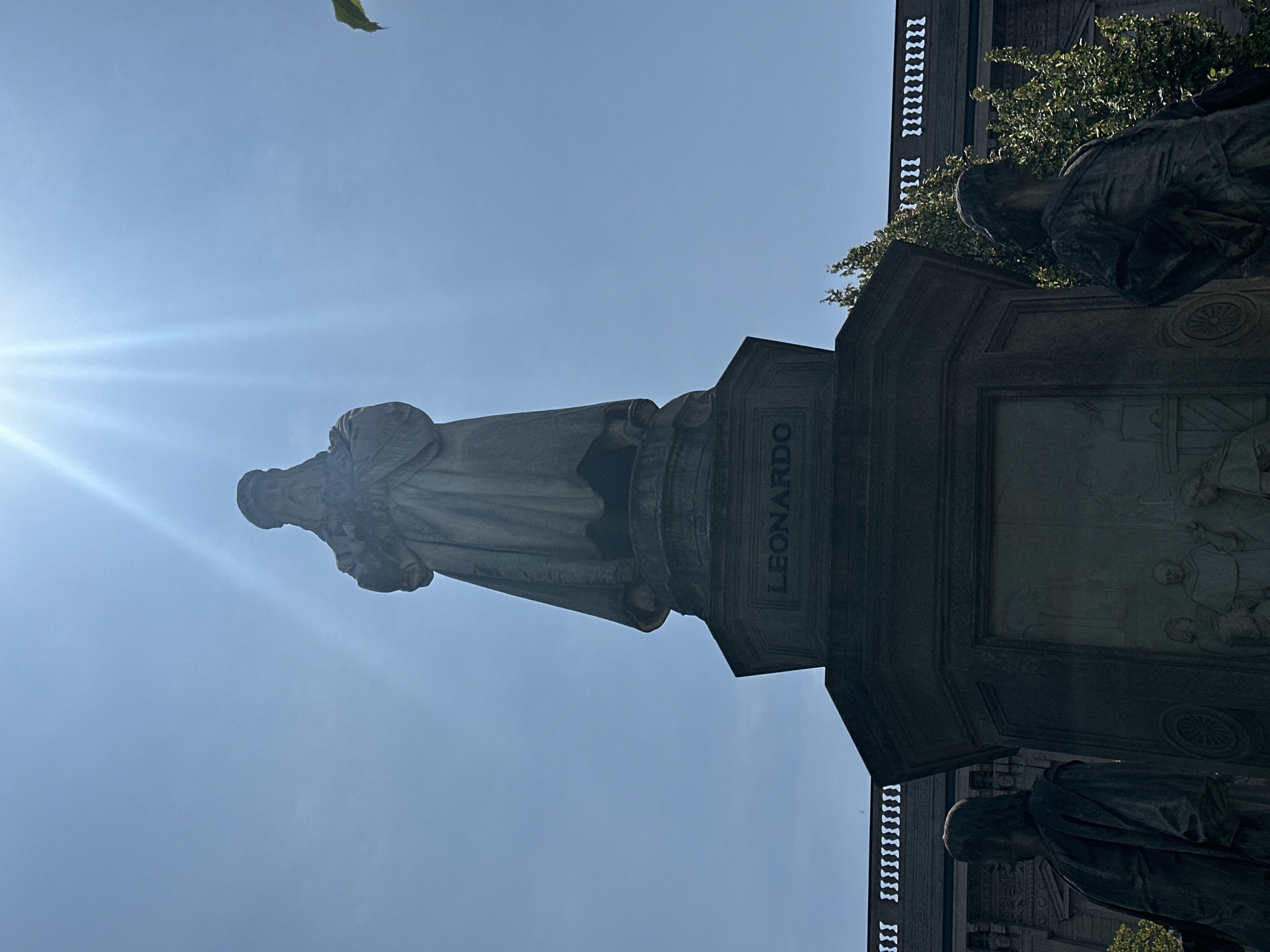 A low-angle, full shot shows the Monument to Leonardo da Vinci in Milan, Italy, a tall stone sculpture dedicated to the Italian Renaissance artist and inventor. The monument is situated in a plaza with trees and a building in the background. The sky is bright blue with the sun shining down, creating lens flares. The monument features a central statue of Leonardo da Vinci, with a beard and flowing robes, holding a book. The base of the monument is adorned with bas-reliefs depicting scenes from his life and work, including figures and machinery. Two smaller statues of students or patrons stand at the base, facing outward. The monument is made of stone and appears to be well-maintained.