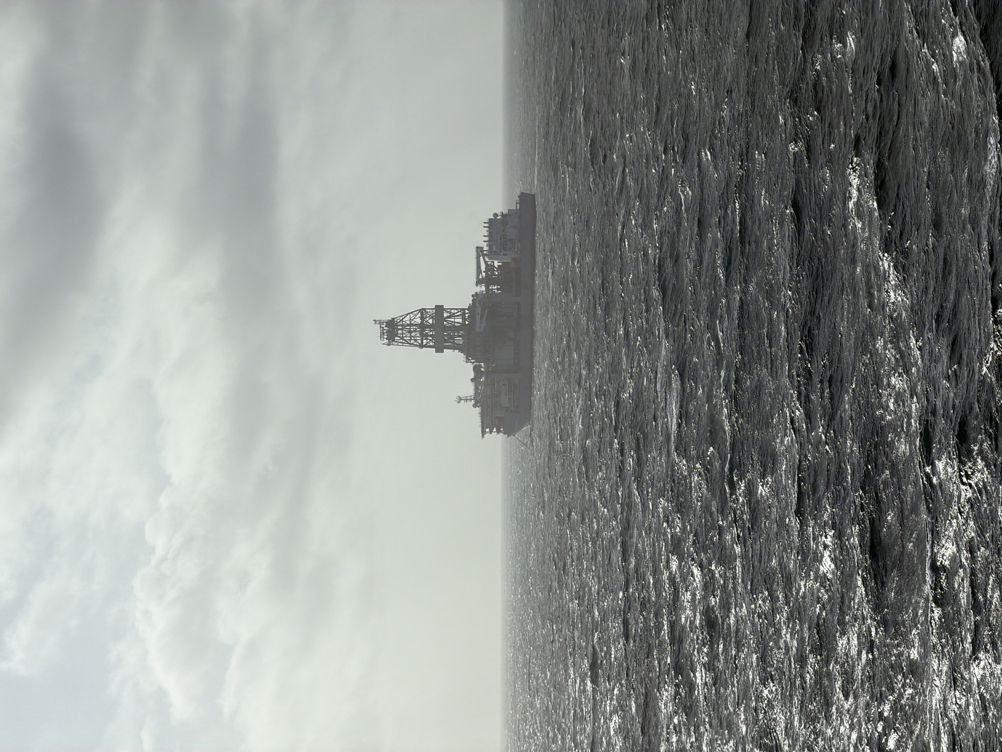 A monochromatic, rotated, long shot depicts an oil rig at sea. The rig, a tall structure with intricate framework, stands against a backdrop of a cloudy sky. The sea occupies the majority of the frame, its surface textured with numerous small waves creating a pattern of light and shadow. The location is not specified in the prompt.