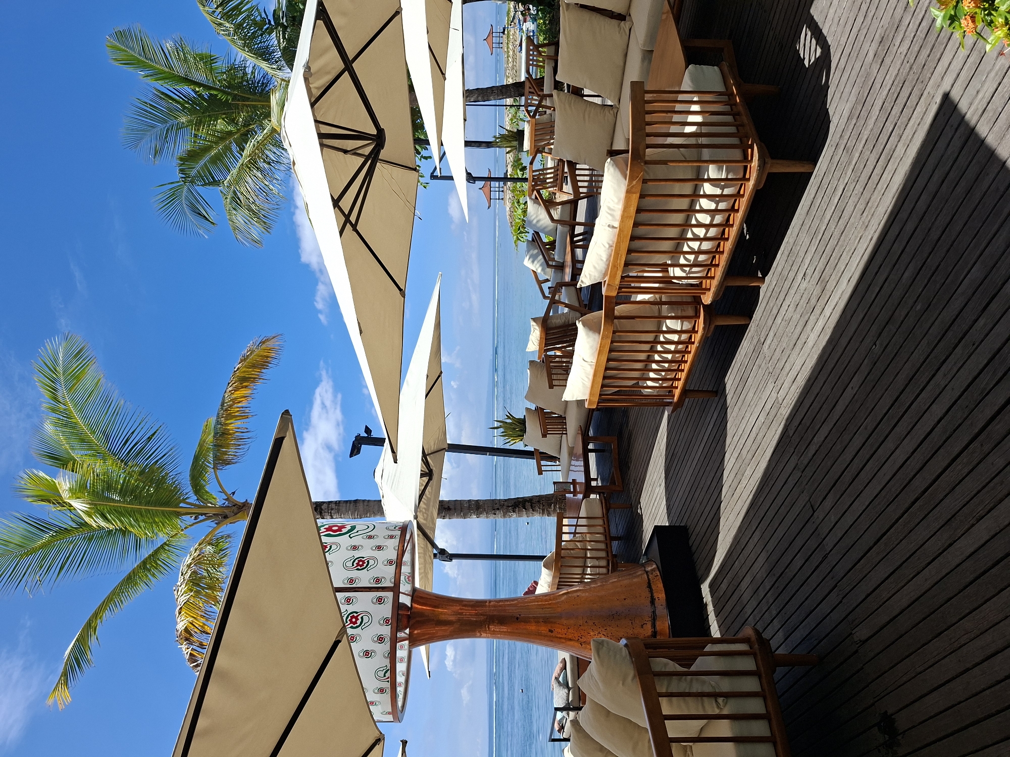 The image depicts an outdoor seating area at a resort or restaurant in Denpasar, Indonesia. The scene is oriented with a beach view in the background. Several white patio umbrellas provide shade for wooden lounge chairs and sofas with light-colored cushions. The seating is arranged on a dark wooden deck. Palm trees are visible, contributing to the tropical atmosphere. The sky is blue with sparse clouds. A large copper or bronze-colored, hourglass-shaped object stands between seating areas, possibly a decorative or architectural feature. People are visible in the distance near the water.