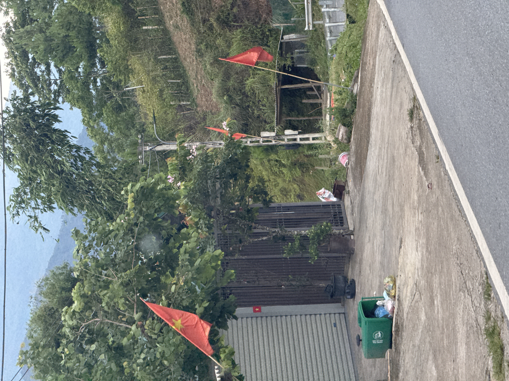 This image depicts a rural or semi-rural scene in Tu Le, Vietnam, characterized by lush green hills and a paved road. In the foreground, a gray asphalt road with a white solid line on its edge transitions to a concrete surface on the left. A green plastic wheelie bin lies on its side on the concrete, spilling multicolored plastic bags of trash. Adjacent to this, a building features a gray corrugated metal roll-down garage door and a dark metal gate. A large Vietnamese flag (red with a yellow star) is flown on a pole partially obscured by trees to the left of the building, and a smaller version of the flag is affixed to the building's wall next to the gate. Several potted plants are positioned near the gates.
Further to the right, a concrete utility pole with power lines and a streetlight fixture stands amidst dense foliage. Two plain red flags are erected on poles further up the sloped terrain. A rudimentary wooden structure with a corrugated metal roof is visible on the hillside behind a dirt path and a simple fence line. The background is dominated by steep, tree-covered mountains under a hazy sky. No individuals or animals are present.