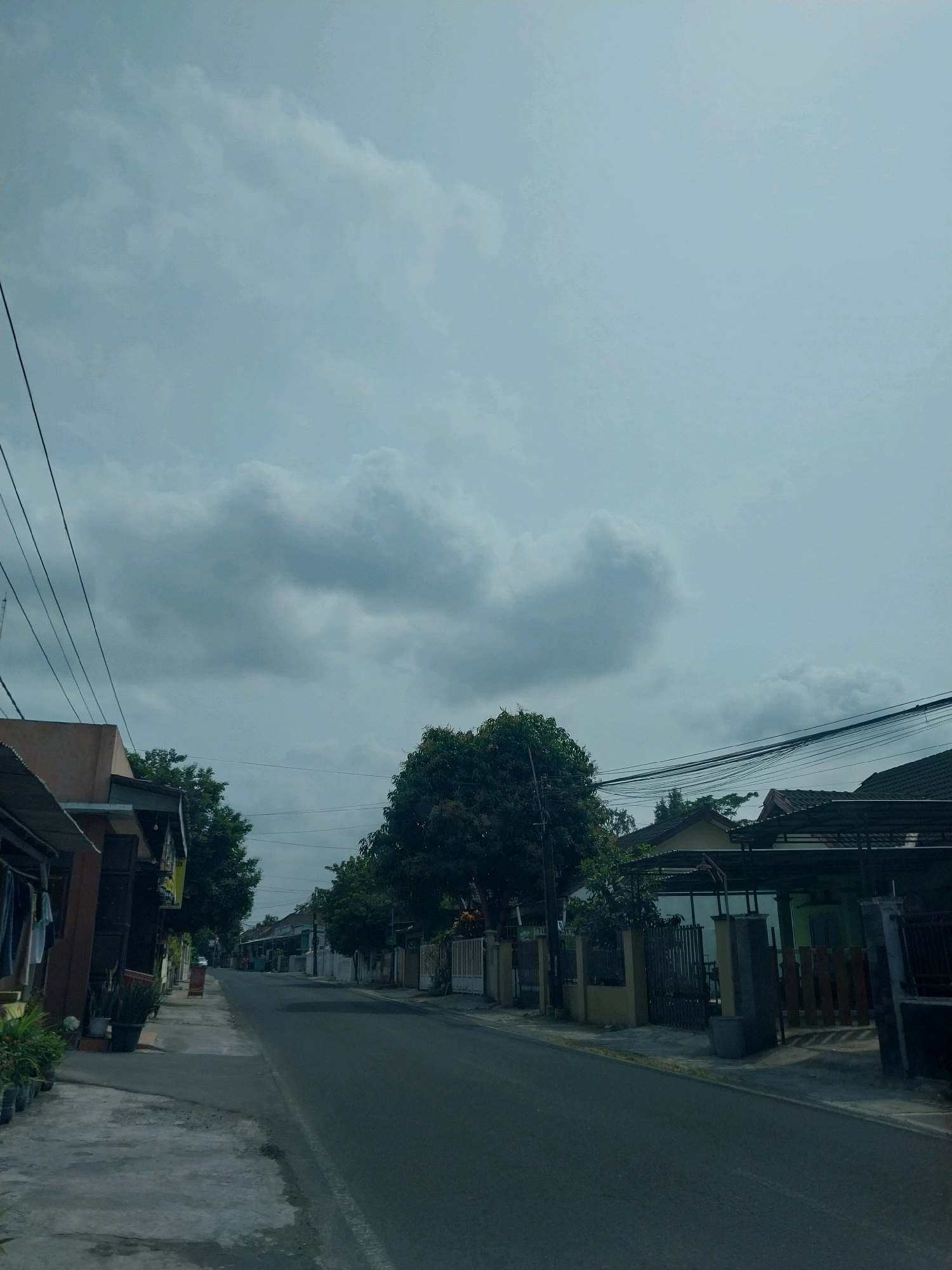 The image depicts a street scene in Purwomartani, Indonesia. The perspective is looking down a paved road lined with houses on both sides. The sky is overcast with thick, gray clouds. Overhead, power lines are strung between poles, creating a network of wires. On the left side of the road, a sidewalk is visible, with houses and some potted plants. Clothing hangs on a rack outside one of the houses. On the right side of the road, more houses with fences and gates are visible, along with a large tree in the middle ground. The overall impression is that of a residential street under a cloudy sky.
