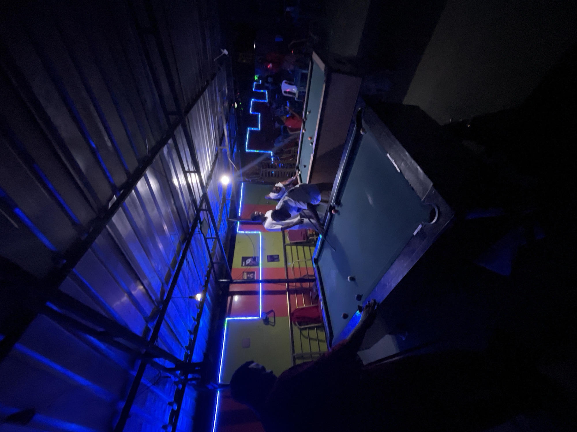 A dimly lit pool hall in Ado, Nigeria, features multiple pool tables with players engaged in games. The hall is illuminated by blue LED strip lights, creating a vibrant, yet slightly dark atmosphere. The walls are painted in a pattern of green, red, and yellow.