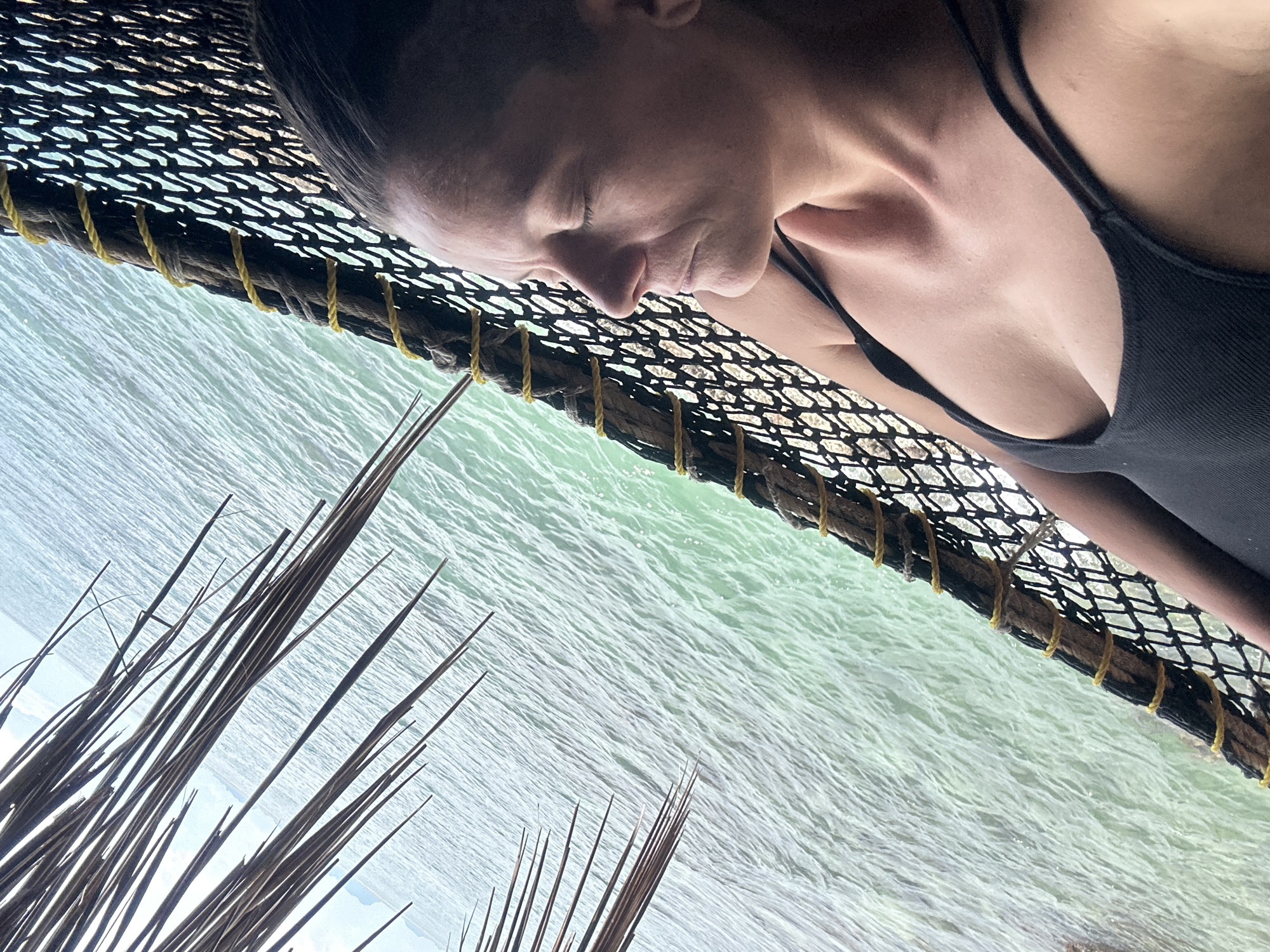 A woman with closed eyes reclines in a hammock against a backdrop of the ocean. The hammock is made of black netting with thick rope and yellow cord accents. Palm fronds are visible in the upper left corner, partially obscuring the sky. The ocean water is a vibrant turquoise with visible ripples. The scene appears to be on Koh Phangan Island, Thailand, likely near a beach or resort.