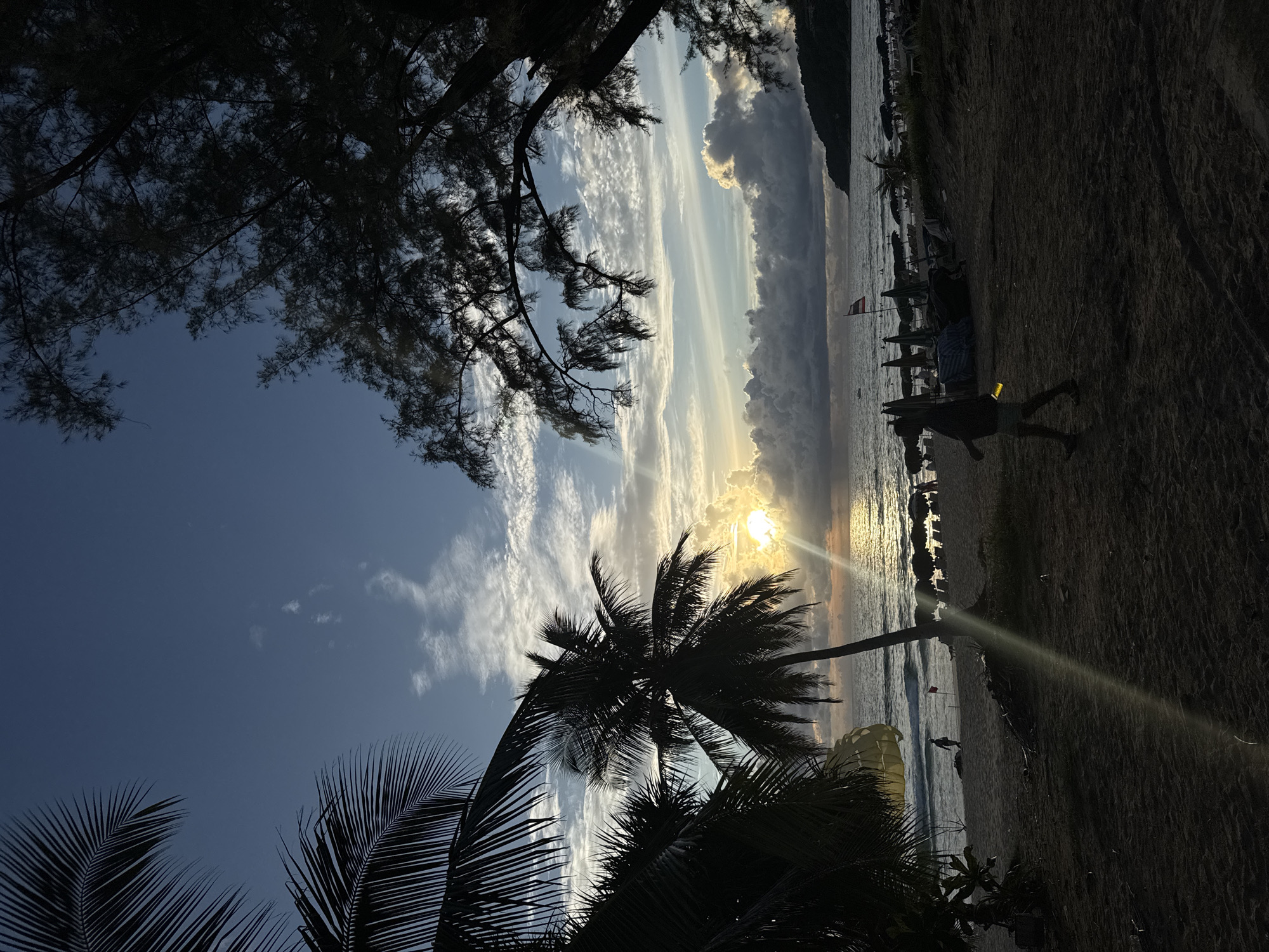 This is a vertical, slightly low-angle shot of a beach in Karon, Thailand, during what appears to be either sunrise or sunset. The sun, partially obscured by clouds, casts a golden glow across the sky and reflects on the calm ocean water. Silhouetted against the vibrant sky are palm trees, with their fronds prominent in the foreground. In the distance, a line of beach umbrellas and loungers suggests a resort or populated area.
The beach itself is sandy, with a few people visible. One person is silhouetted as they walk along the shoreline, and another appears to be running. Further back, near the water's edge, a yellow parasail is visible, suggesting recreational activities. A single small flag with red and white stripes stands planted on the beach. The overall mood is serene and tropical, with the warm light creating a peaceful atmosphere. The sky is a mix of blue and dramatic clouds, some illuminated by the sun, others darker and more textured. The natural beauty of the scene is emphasized by the silhouetted tropical foliage.