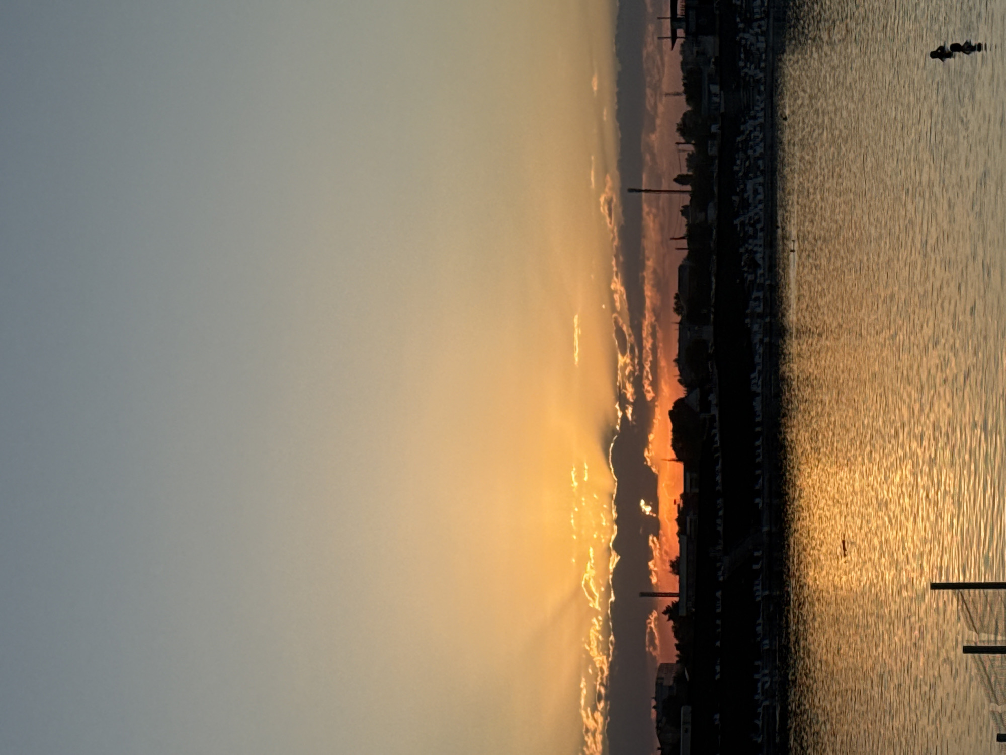 This is a vertical, medium-shot photograph of a sunset over a body of water, likely a lake or river, in Sol-Iletsk, Russia. The sky transitions from a light blue at the top to a gradient of orange and yellow near the horizon, where the sun is partially obscured by clouds. Rays of sunlight emanate from behind the clouds, casting a warm glow.
In the background, a silhouetted cityscape runs along the edge of the water. Visible elements include buildings, trees, and industrial structures such as smokestacks or towers. A line of empty chairs or loungers is visible along a raised embankment in front of the cityscape, suggesting a public beach or recreational area.
The water in the foreground reflects the colors of the sunset, creating a shimmering, golden surface. Several small, dark shapes are visible in the water, appearing to be people swimming. One figure is clearly visible in the lower right corner, with only their head and shoulders above the water. Another dark shape further in the distance may be an animal, such as a duck or goose, or another person. In the lower left of the frame, a portion of a fence or railing is visible, suggesting a man-made structure or boundary near the water's edge.
