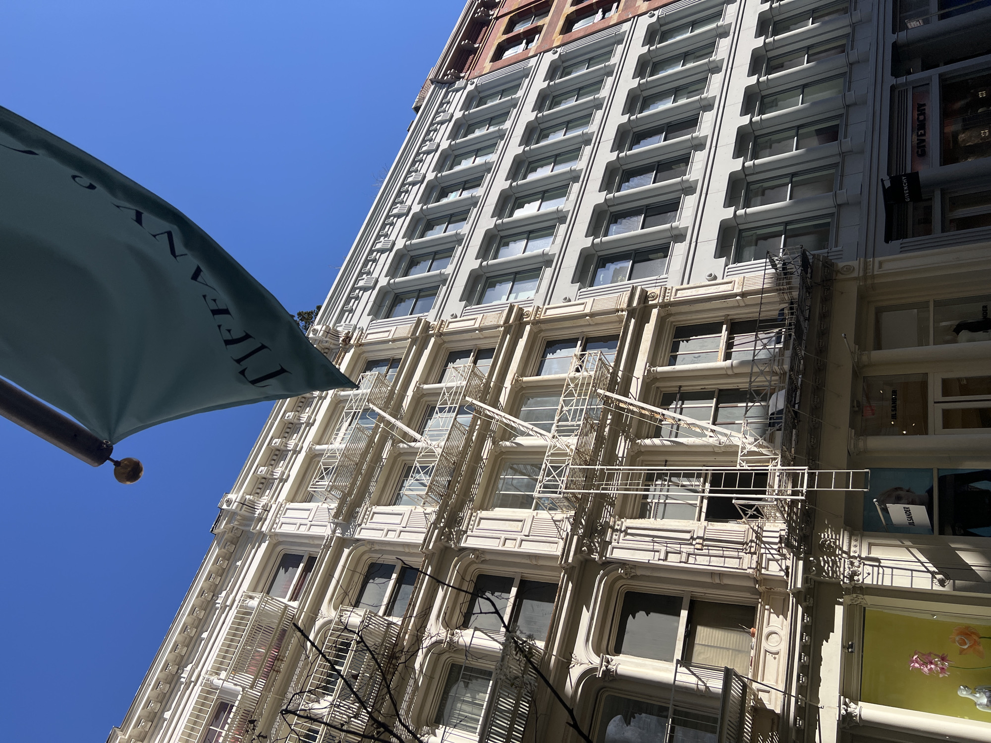 Captured from a low-angle perspective, the image portrays a tall building facade with an ornate, layered architectural style in New York City, United States of America. The building has a distinctive combination of off-white and light gray hues, featuring rows of uniform windows across multiple stories. The facade is adorned with decorative molding, vertical columns, and fire escapes extending along the side. A flag with the word "Tiffany" is partially visible on the left side of the frame, hinting at the building's potential location or association. To the right of the primary building is a structure with visible storefronts bearing names like "GIVENCHY" and "JIL SANDER," indicating a commercial area. The sky, a clear blue, provides a backdrop to the building's structure.