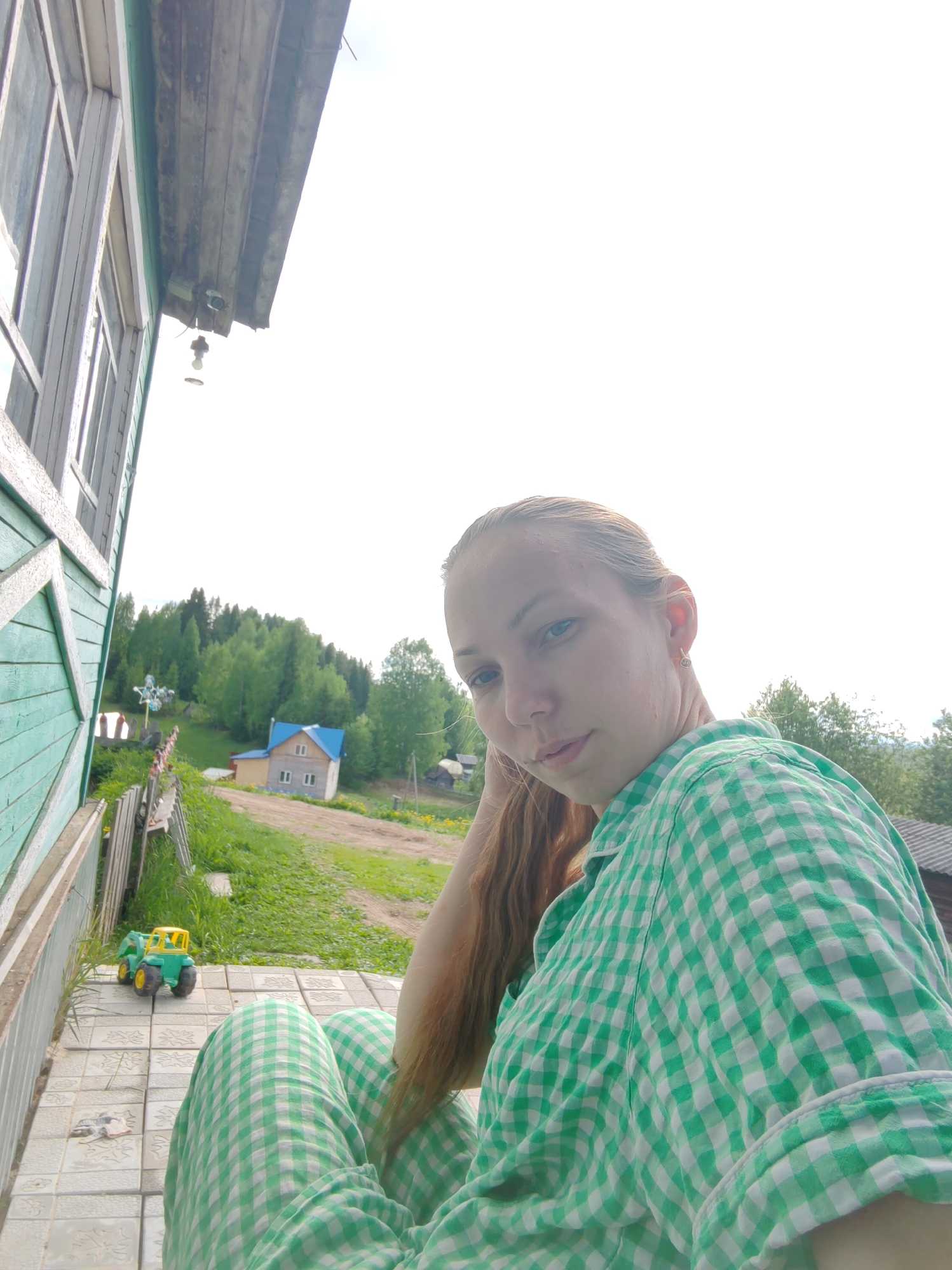 The image shows a woman in a green and white checkered shirt, possibly pajamas, seated on a tiled porch. Beside her is a green and yellow toy tractor. In the background is a rural scene, including a house with a blue roof, a grassy area, and trees. The location is likely in the city of Чухлэм, Russia.
