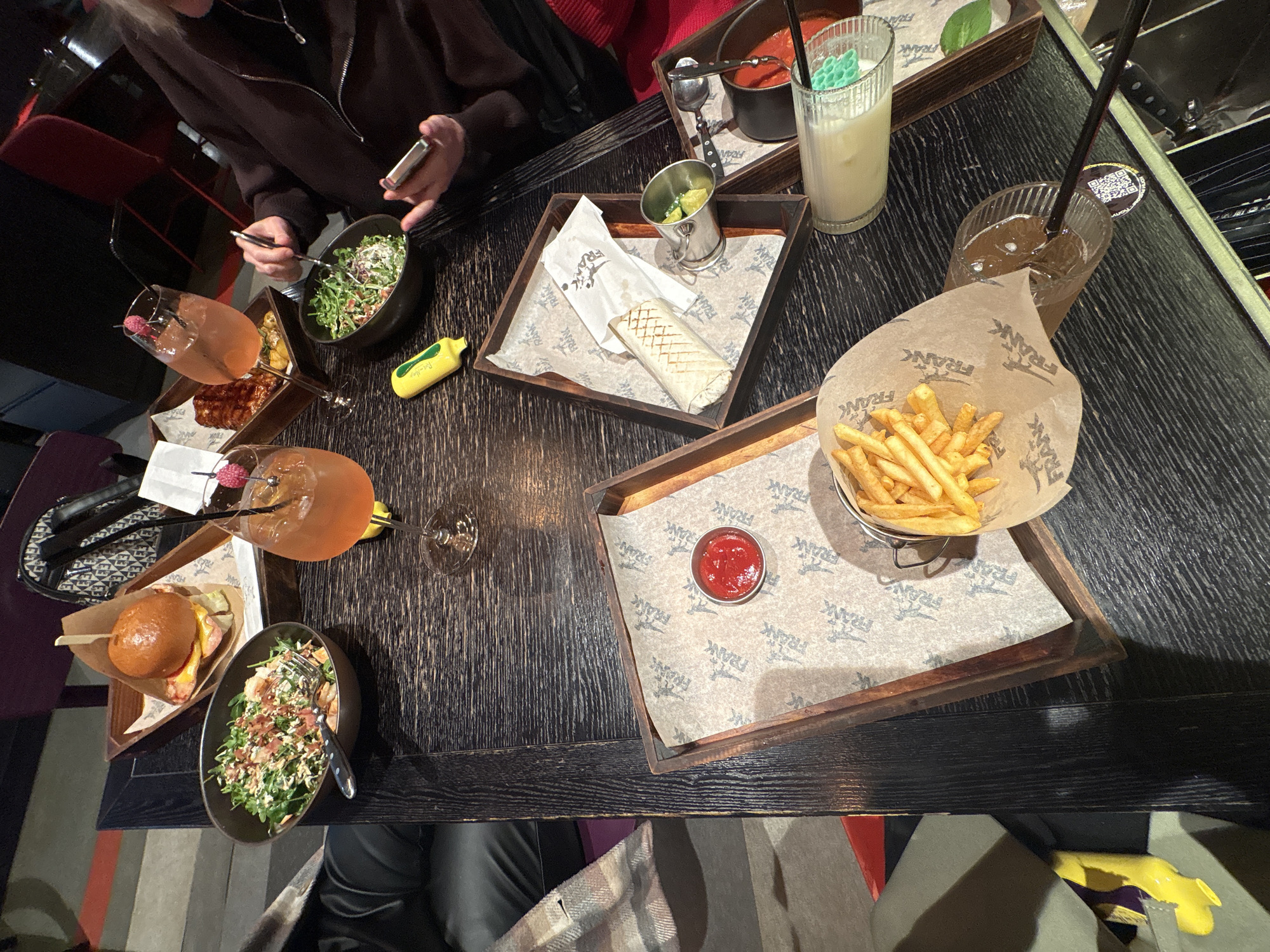 Captured from a high-angle perspective, the image showcases a black wooden table laden with a variety of foods and beverages. The scene appears to be in a restaurant in Saint Petersburg, Russia, as the brand "FRANK" is displayed on the paper liners of the trays.
At the table are at least two individuals, one seen using a smartphone and eating a salad with a fork, while the other is only partially visible. Various dishes and drinks are arranged on the table:
- A burger and salad on a tray, with a drink nearby.
- A tray featuring a wrap and a small container of pickles.
- A container of french fries, accompanied by a small dish of ketchup on a separate tray.
- A cocktail with a raspberry garnish.
- A tall, light-colored drink with a decorative green ornament.
- A small pot of tomato sauce with a spoon.
- A glass of iced tea with a black straw.
- A small, yellow, toy-like object is placed near one of the salads.
The table is situated near other dining areas, hinted by the presence of chairs and a glimpse of a purse in the periphery. The lighting in the restaurant is dim, enhancing the visual appeal of the food and drinks on the table. The overall ambiance suggests a social gathering centered around a meal.