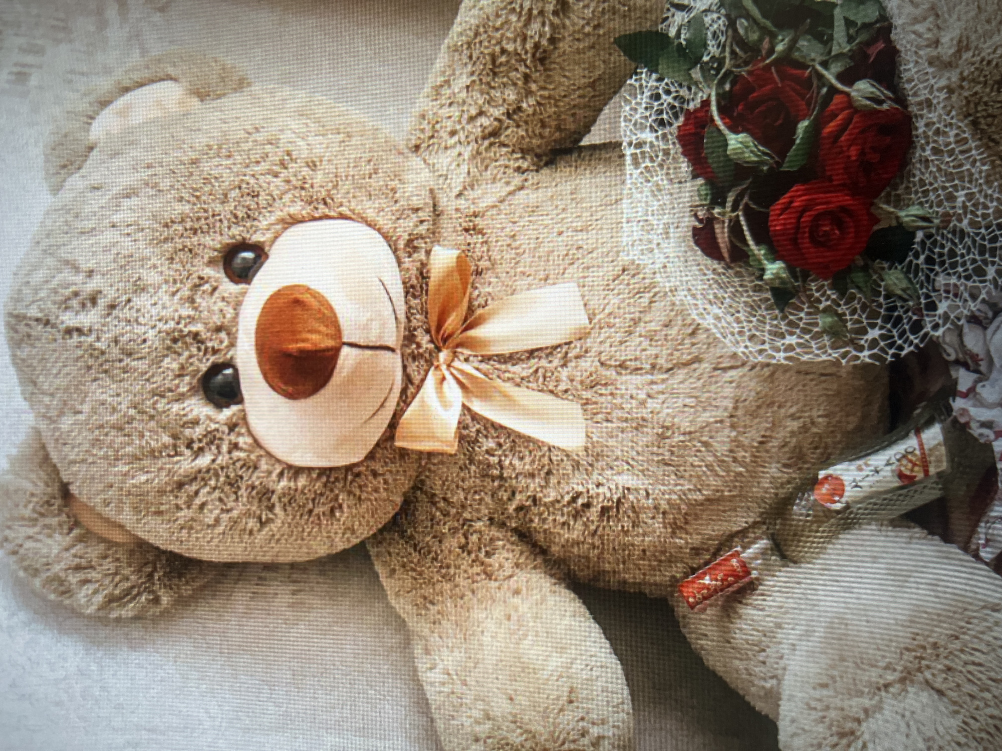 A large, light brown, plush teddy bear is the primary subject of the image, seated upright. It wears a light gold satin bow around its neck. In front of the teddy bear, to its left, rests a small, clear glass bottle with a red label and a red cap. To the right of the bottle and in front of the teddy bear's lower body is a small bouquet of red roses, partially wrapped in white netting. The background consists of a textured, light-colored surface, possibly a wall or fabric. The lighting suggests an indoor setting, with soft shadows. The image does not provide any information about the city of Moscow, Russia.