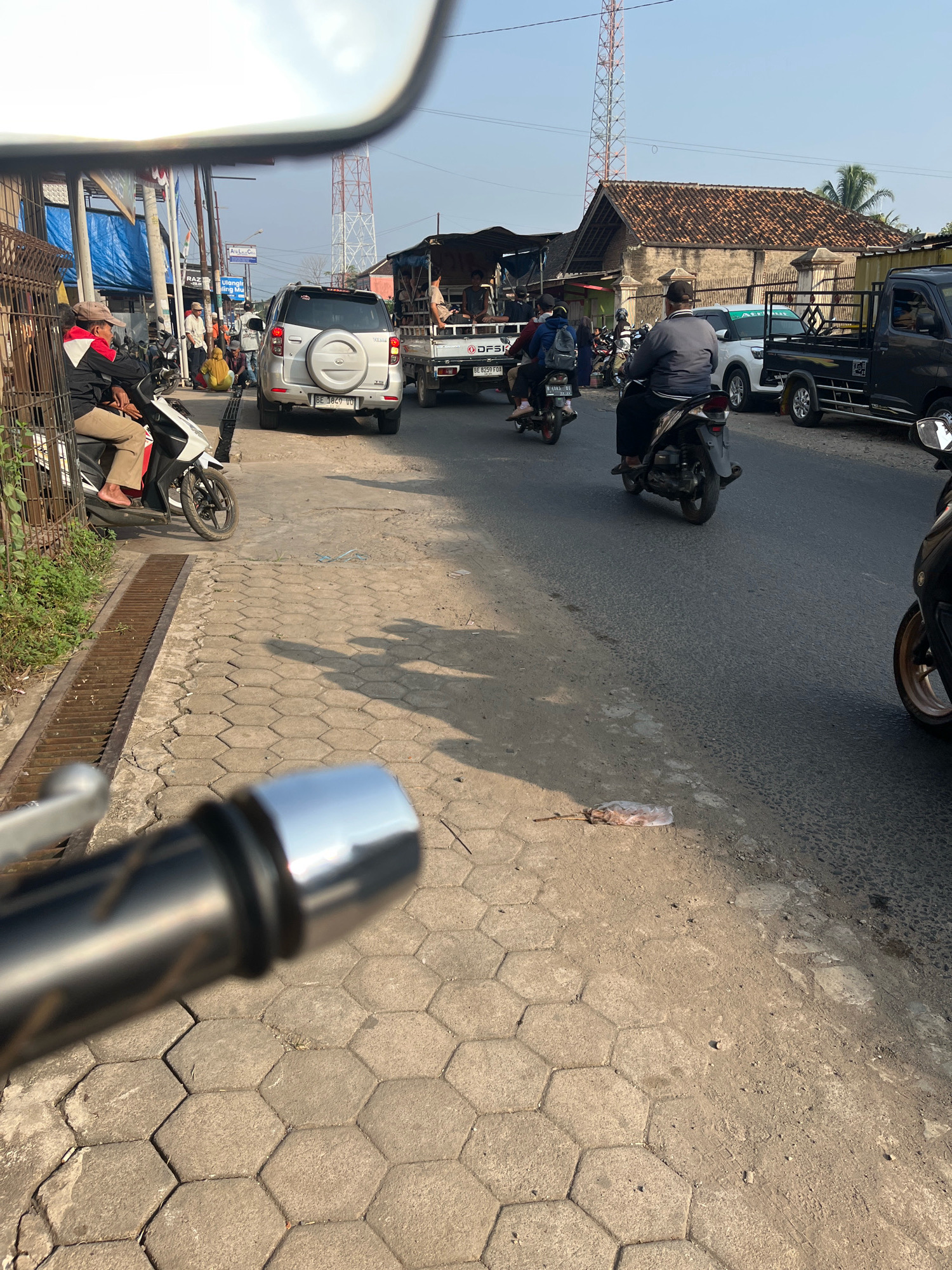 The image captures a street scene in Kemuning, Indonesia, likely during the daytime, given the clear sky and shadows. In the foreground, the handlebars and a portion of the front of a motorcycle are visible, suggesting the photo is taken from the perspective of a rider.
The road is paved with asphalt, and to the left, a pedestrian walkway is made of hexagonal concrete paving stones, bordered by a metal grate drain. Along the edge of the walkway, there is a low metal fence and some greenery.
Several vehicles and individuals are present on the street. A white SUV with a spare tire mounted on the back is parked or moving slowly. A black pickup truck is also visible. Several motorcycles are in motion or parked. One person is seated on a motorcycle in the foreground, to the left, wearing a red and black jacket and beige pants. Another person is riding a motorcycle further down the road, wearing a grey jacket and black pants, with a black cap. A motorcycle with two riders is also visible.
In the background, there are buildings with tiled roofs, some with weathered facades. A prominent red and white transmission tower stands tall against the blue sky. There are also power lines and streetlights. The general impression is of a busy, semi-urban or peri-urban area with a mix of residential and commercial elements. There appears to be some debris on the ground near the edge of the road, including a plastic bag.