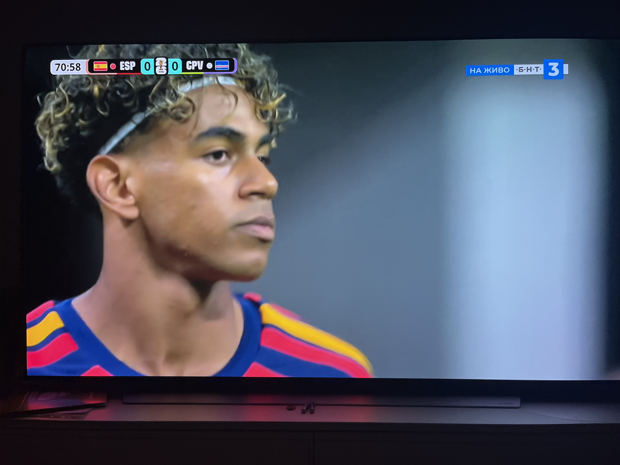 The image shows a close-up of a television screen displaying a football match. The score is 0-0 between ESP and CPV at 70:58. A male player with curly hair and a white headband is visible on the left side of the screen, wearing a blue shirt with red and yellow stripes. The right side of the screen displays a blue banner with white text that reads "НА ЖИВО Б.Н.Т 3", indicating a live broadcast on BNT 3, a Bulgarian national television channel. The television is placed on a dark piece of furniture. The image appears to be taken in a dimly lit room. There is no discernible location in the city of Varna, Bulgaria.