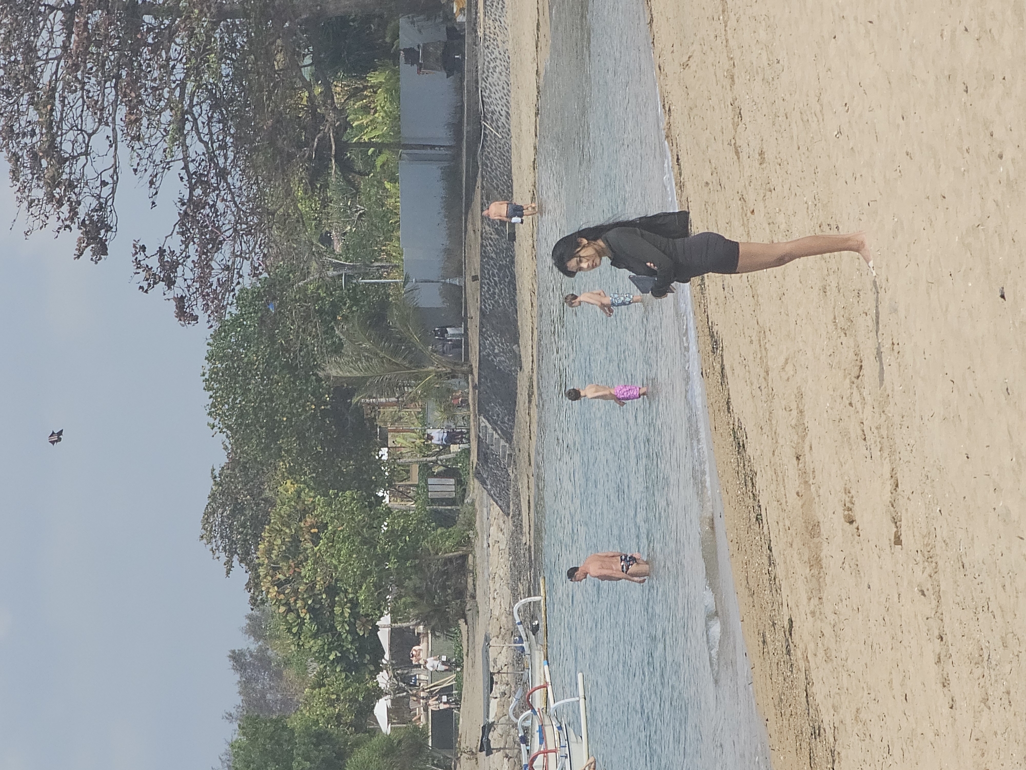 A low-angle, eye-level, full outdoor shot captures a diverse group of people at a tropical beach in Denpasar, Indonesia. In the foreground, a young woman with long dark hair stands on the sandy beach, facing the water with her arms crossed. She is wearing a black long-sleeved rash guard and black shorts. Several individuals are visible in the shallow, clear blue water. A man with dark hair and patterned swim trunks stands waist-deep, looking to his left. To his right, a young boy in pink swim trunks stands chest-deep, looking towards the beach. Further out, another young boy in patterned swim trunks is also wading in the water. In the background, there are several traditional outrigger boats pulled onto the sand. Behind the boats, a sandy embankment leads up to a developed area with lounge chairs and umbrellas, suggesting a resort or public beach access. A stone wall and lush green trees form a backdrop. In the sky, a small kite is visible. The overall scene depicts a relaxed day at the beach.