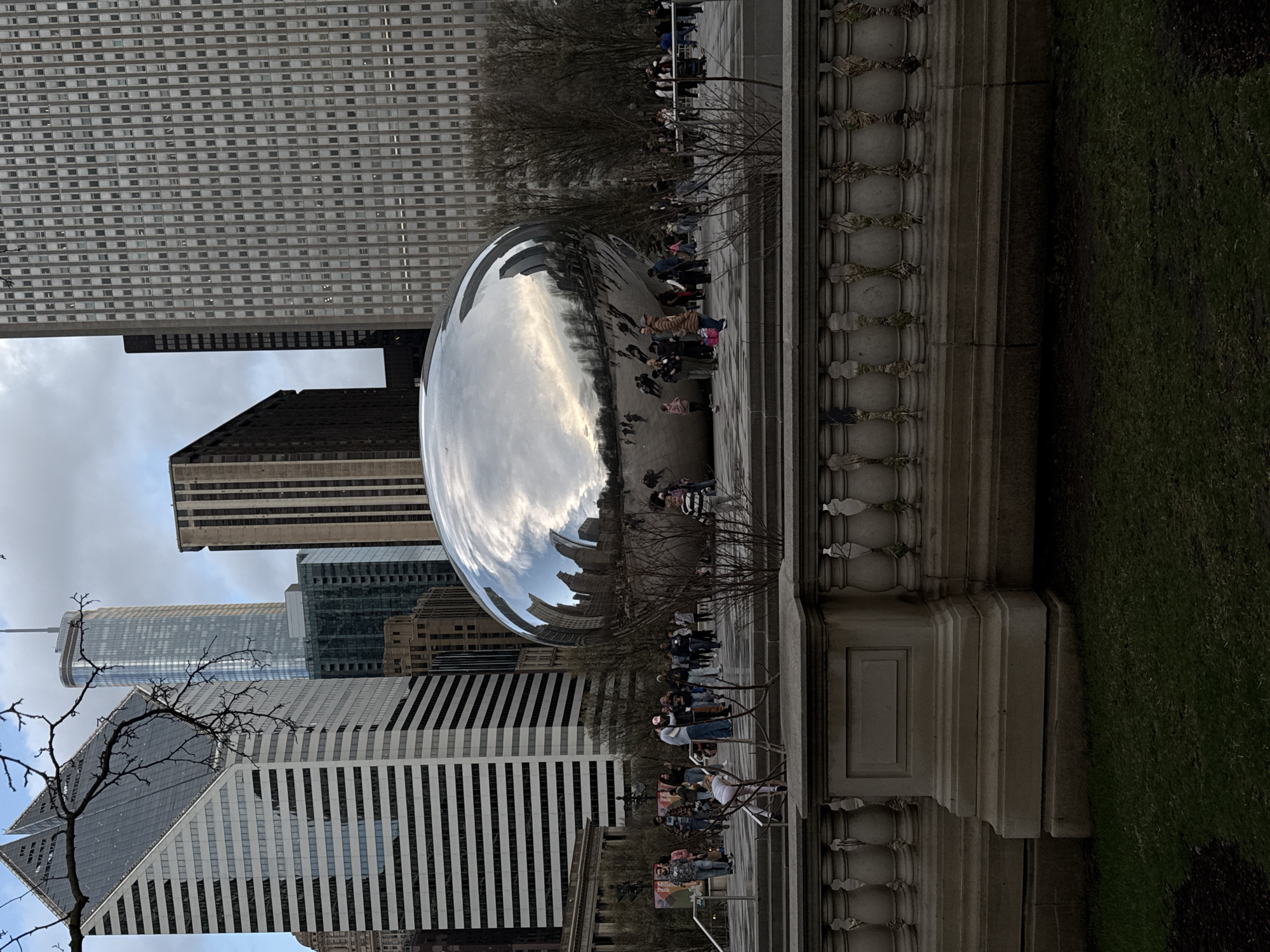 The image captures a scene in Chicago, Illinois, USA, featuring Cloud Gate (commonly referred to as "The Bean"), a popular public art sculpture located in Millennium Park. A crowd of people is interacting with the sculpture, some walking around it, others standing close for photos. The sculpture's reflective surface mirrors the surrounding urban environment, including various skyscrapers of differing heights and architectural styles. A decorative stone barrier is situated on the right, separating the paved area from a patch of green lawn. The weather appears overcast, as suggested by the sky visible in the reflections and the overall lighting. A bare tree is positioned on the left, its branches reaching toward the buildings.