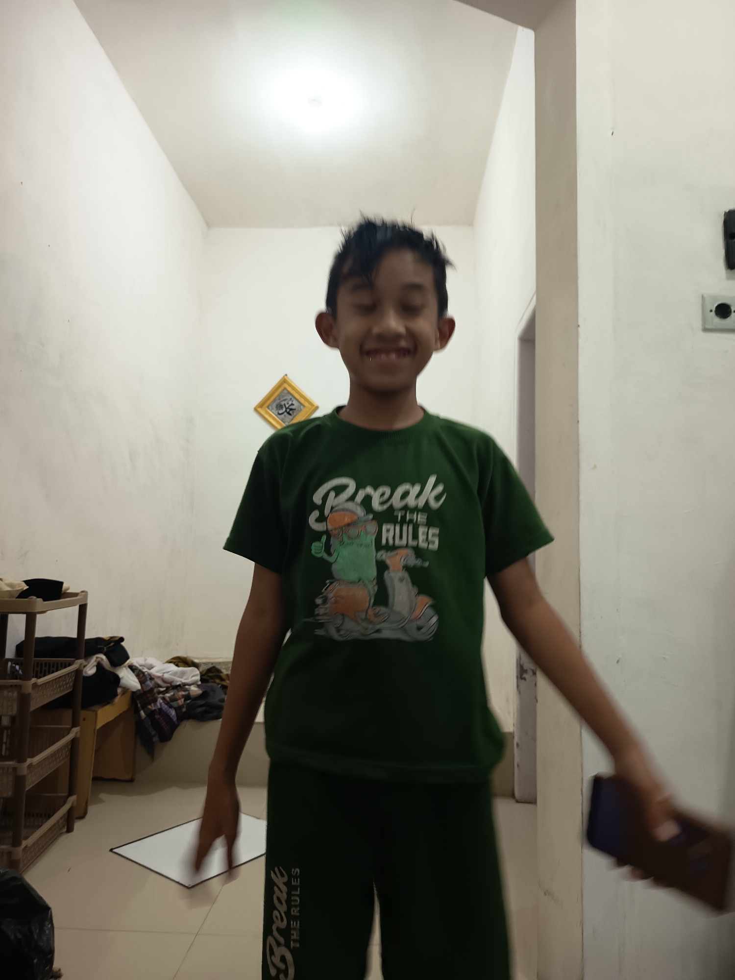 The image depicts a young boy standing indoors, likely in a residential setting in Lemahbang, Indonesia. He is wearing a dark green t-shirt with the text "Break the Rules" and a graphic of a cartoon character riding a scooter. He is also wearing matching dark green pants. The boy is smiling and has his mouth open, with his teeth visible. His hair is styled upwards.
The background shows a narrow room with white walls. On the left side of the image, there is a multi-tiered shelf unit with various items, including clothing and what appears to be a black plastic bag at the bottom. A small framed picture with Islamic calligraphy is hanging on the wall. The floor is tiled. The lighting in the room appears to be from an overhead source. A portion of another person's hand holding a dark-colored rectangular object, possibly a phone, is visible on the right side of the frame, indicating a potential interaction or the presence of another individual just out of view. The overall scene suggests a casual, domestic environment.