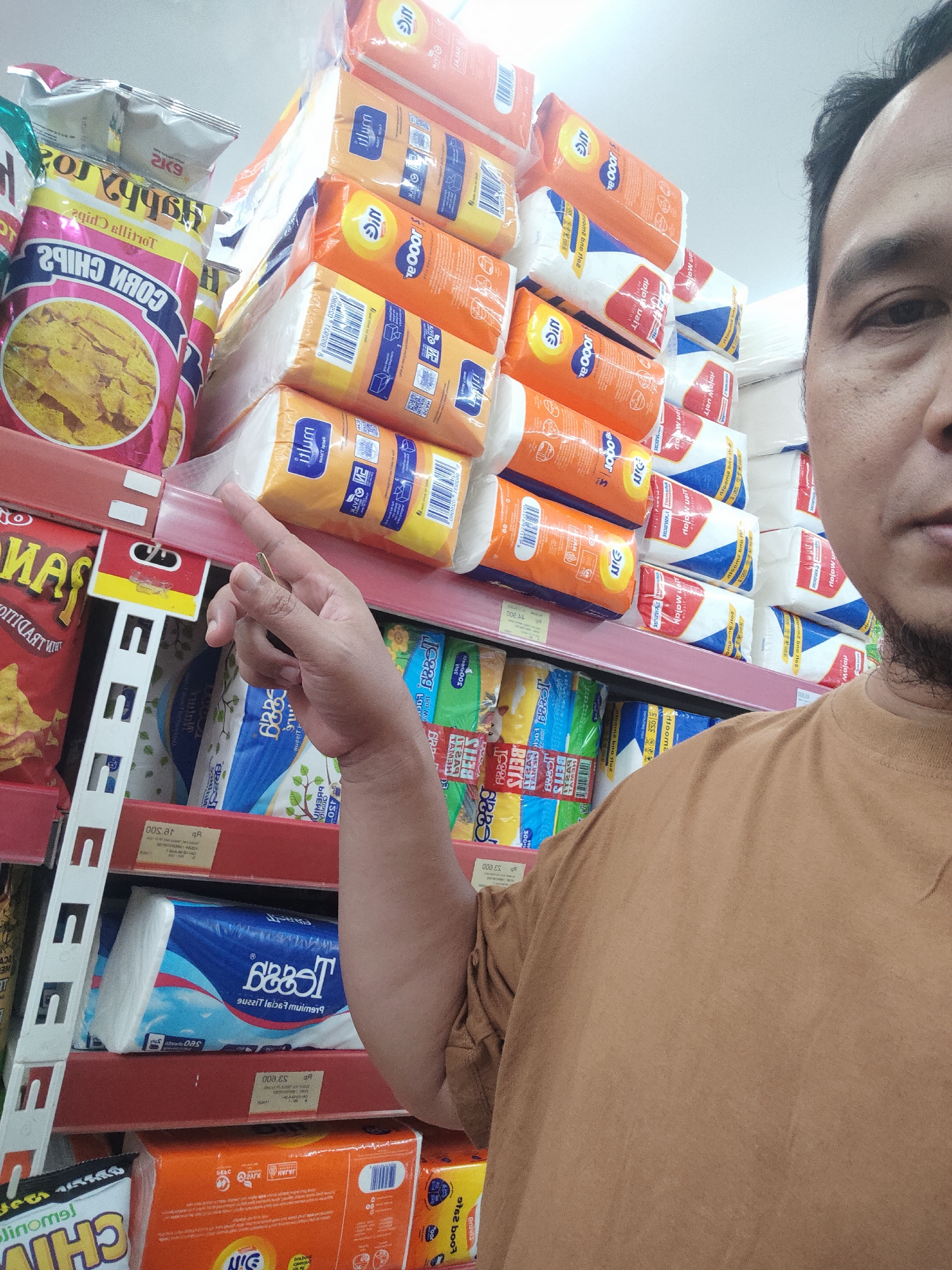 A man in a brown shirt stands in an aisle of a store, holding his right index finger up, pointing towards a shelf stacked with packaged goods, likely toiletries or cleaning supplies. The shelves are organized and filled with various items, including paper towels and what appear to be snack bags. The store appears to be a supermarket or convenience store, possibly in Desa Banyumas, Indonesia, given the context. The man's face is partially visible on the right side of the frame, and he has a beard. The lighting suggests an indoor environment.