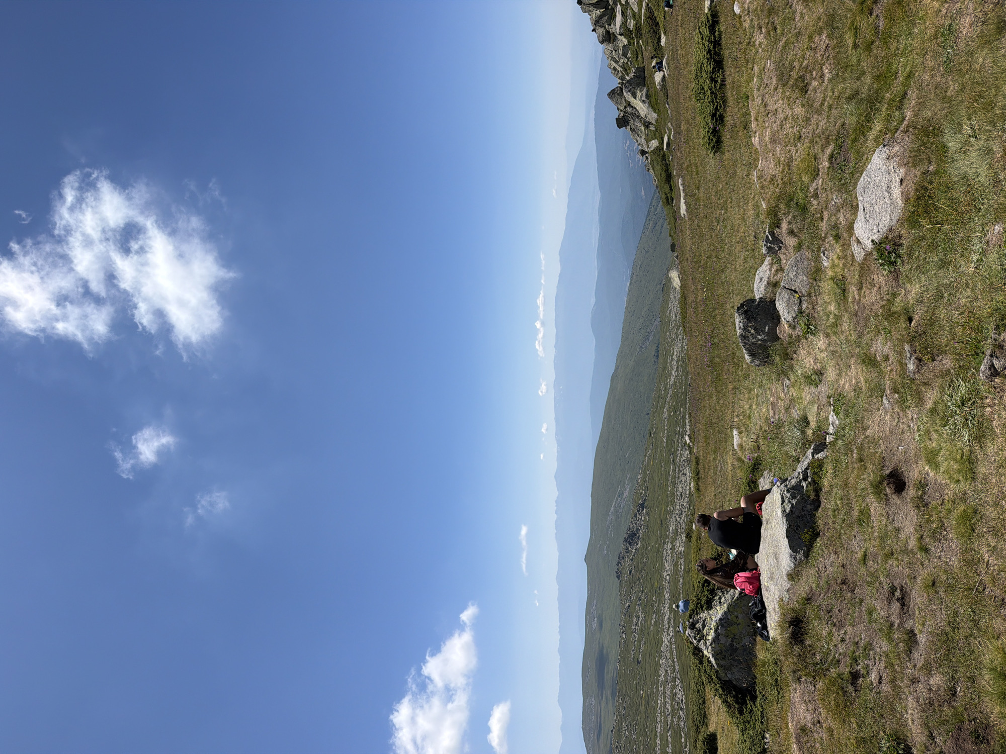 A vertical shot captures a panoramic view of a mountainous landscape under a bright blue sky with scattered white clouds. In the foreground, a grassy meadow is dotted with rocks and patches of dry grass. Two individuals are seated on a large rock in the lower left quadrant of the image. One person, appearing to be female with long brown hair, is wearing a dark top and shorts. Beside her sits another individual, possibly male, wearing a black tank top and shorts. A pink backpack rests on the rock between them. In the middle ground, rolling green hills extend towards a range of hazy blue mountains in the distance. The far background is dominated by a soft blue sky. The scene is indicative of an outdoor recreational area, likely a hiking trail in the Vitosha Mountain region near Bistritsa, Bulgaria.