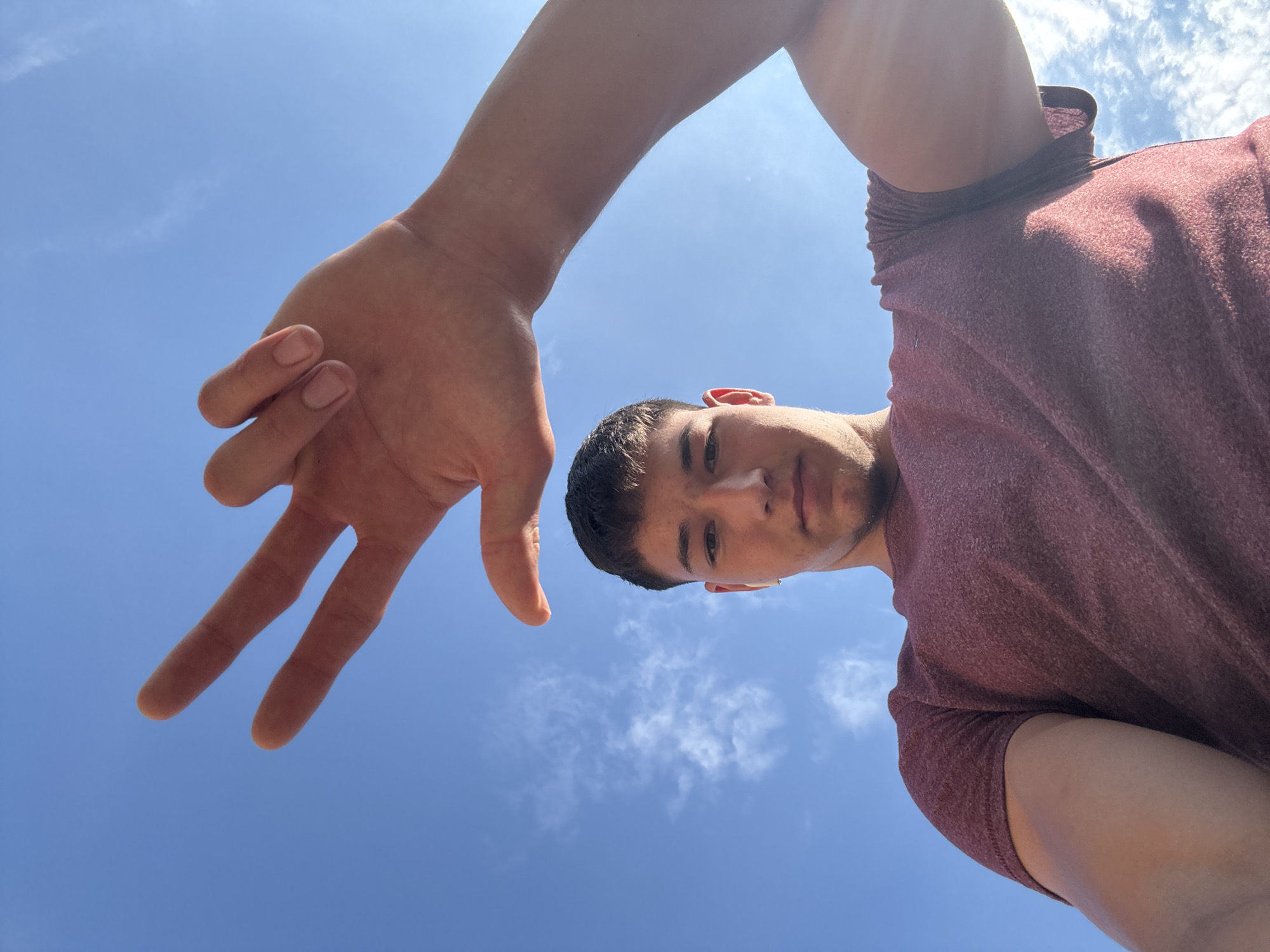 The image shows a low-angle, close-up shot of a young man against a bright blue sky with scattered white clouds. His face is partially visible, looking upwards towards the camera, with a small amount of sunscreen on his nose. He is wearing a maroon, textured t-shirt. In the foreground, his left arm and hand are extended towards the camera, with his fingers spread open. The perspective suggests he is holding the camera or phone to take a selfie, possibly outdoors on a sunny day. The overall impression is casual and carefree, with the bright sky and open hand creating a sense of freedom and a warm atmosphere. No text is visible in the image. The location context is Zlatograd, Bulgaria.