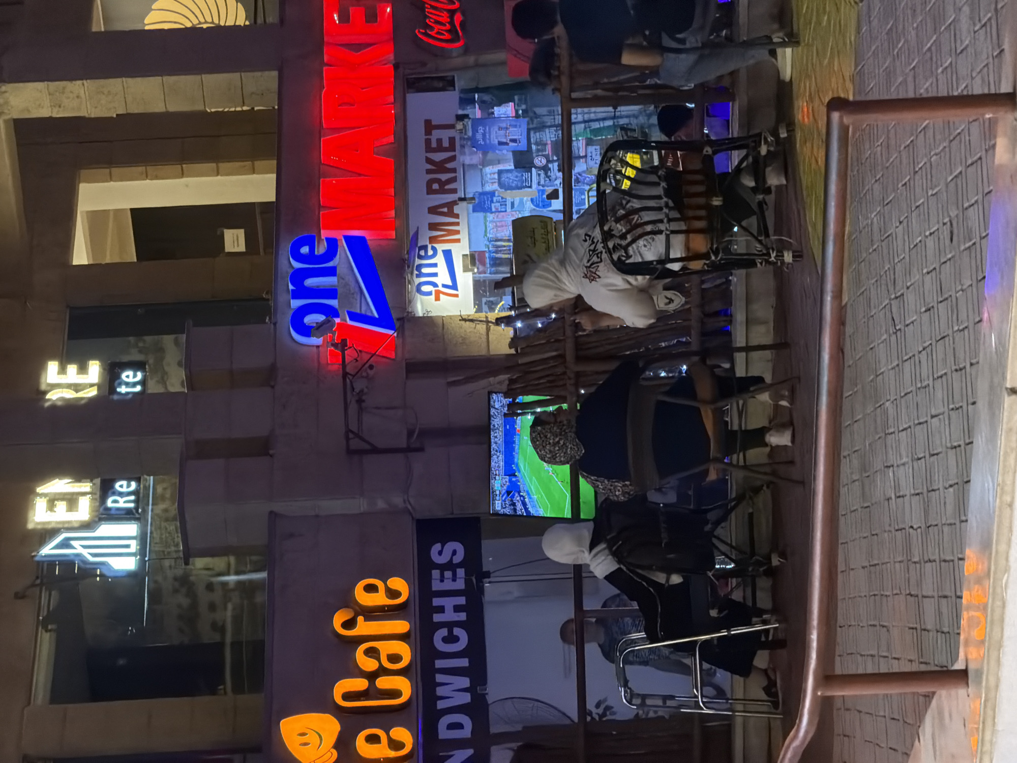 The image shows a street scene at night in Sharm El Sheikh, Egypt. In the foreground, a metal railing runs horizontally, suggesting a walkway or balcony. Beyond the railing, people are seated at tables, some appearing to watch a television screen displaying a soccer game. Several neon signs are visible, including "e Cafe," "SANDWICHES," and "one MARKET" in both red and blue neon. A Coca-Cola logo is also illuminated in red neon. The architecture features stone facades and upper-level windows. The lighting is primarily from the neon signs and ambient streetlights, creating a vibrant, nighttime atmosphere. The people are casually dressed, and the overall impression is of an outdoor seating area of a cafe or market.