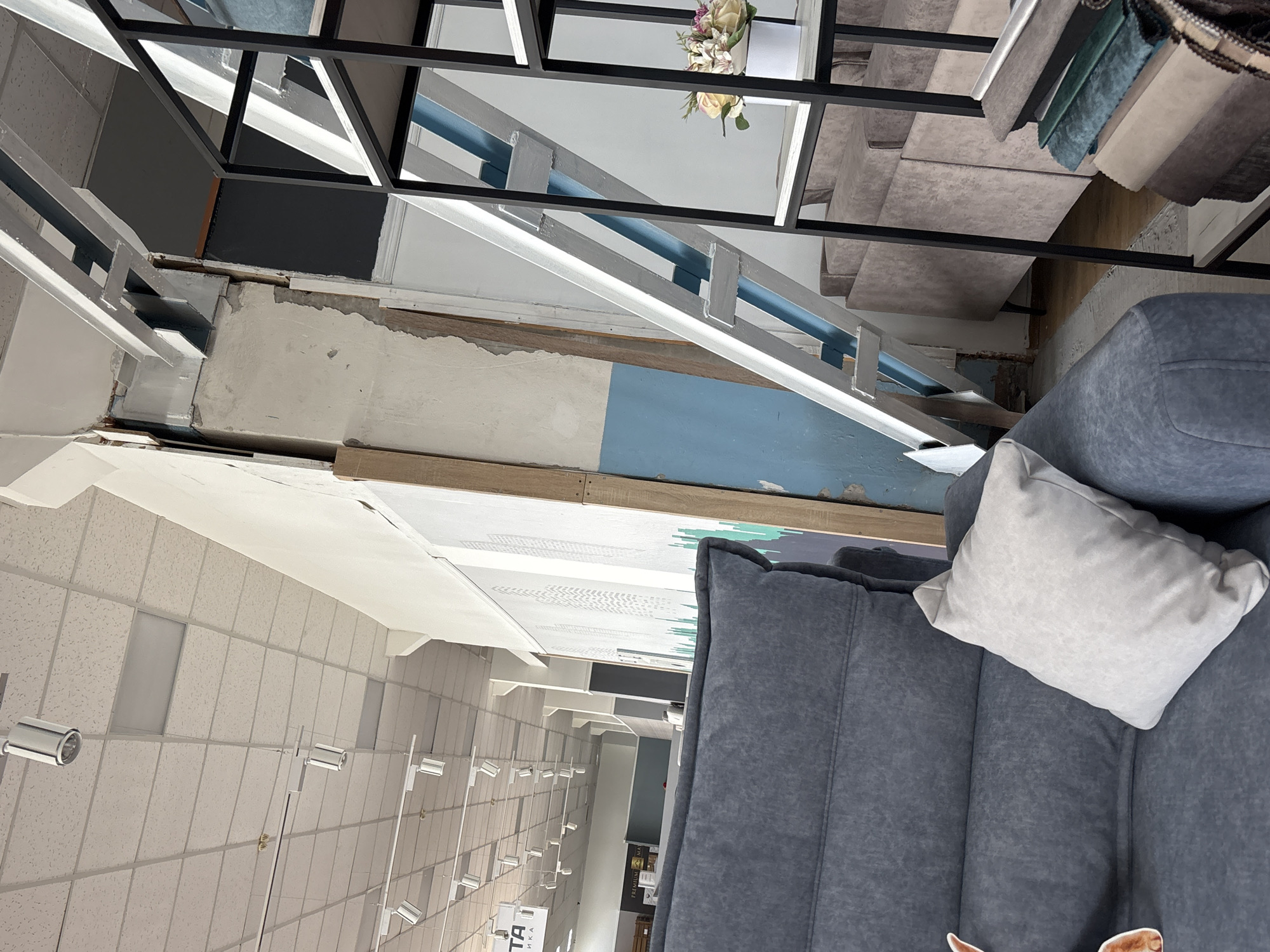 The image depicts the interior of a furniture store, likely in the city of Zaporizhzhia, Ukraine. In the foreground, a section of a large, dark grey sofa with visible stitching and a light grey decorative pillow is prominent. To the right, a black metal shelving unit stands, displaying a bouquet of artificial flowers in a white pot and several folded fabric swatches of various colors and textures on its lower shelves. In the midground, several upholstered furniture pieces, appearing to be sofas, are arranged. The background reveals a commercial space with a dropped ceiling, track lighting, and structural elements of the building, including exposed beams and a partially exposed wall with blue and off-white paint. The overall impression is one of a retail environment focused on home furnishings.