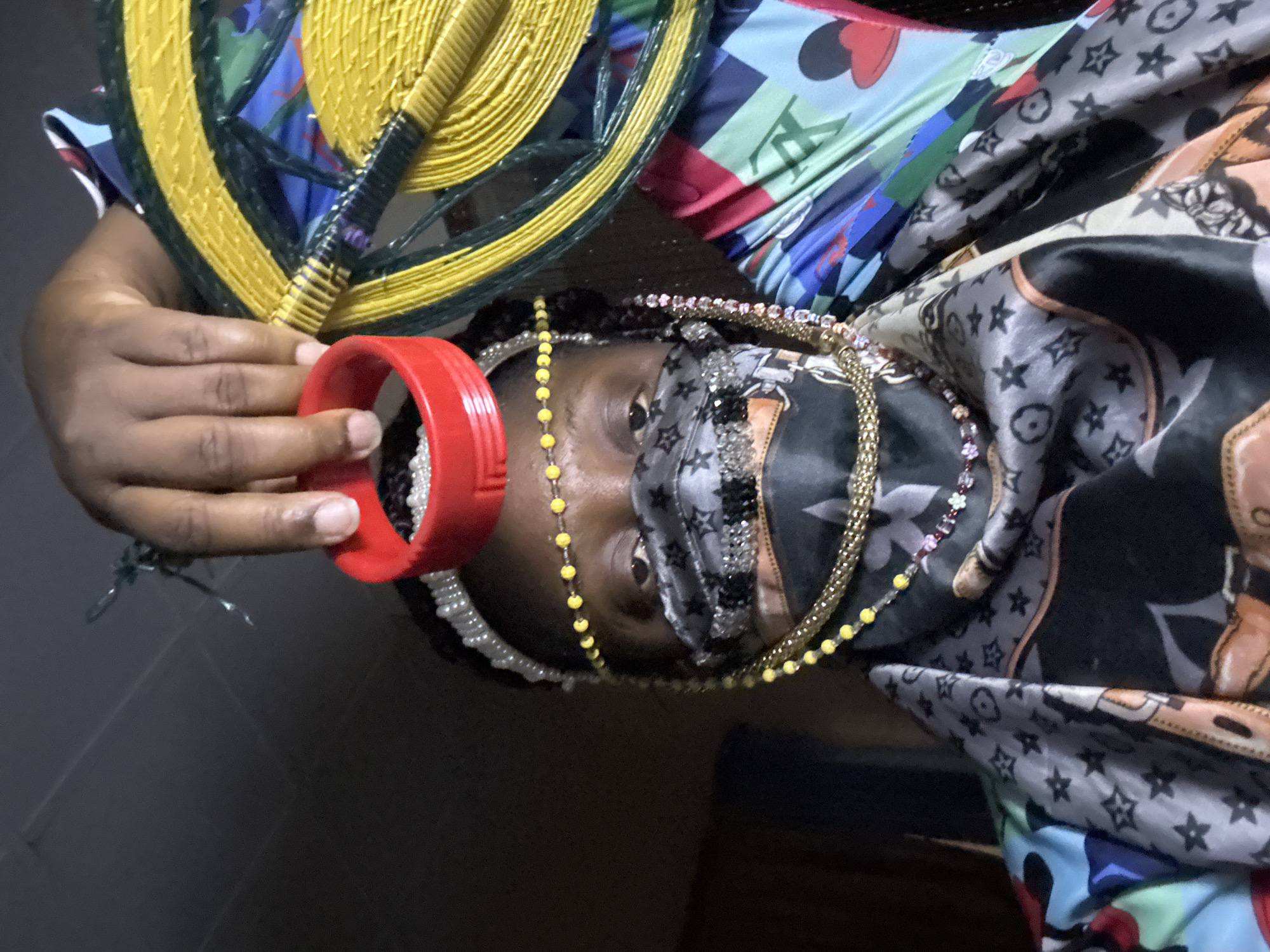 An individual with dark skin and multiple necklaces is partially obscured by a colorful, patterned headscarf. They wear a face mask and hold a red plastic ring near their face, with a woven hand fan featuring yellow and green details also in the frame. The scene appears to be indoors, with a dark floor visible in the background. It is not possible to determine the location of the image within the city of Madangala, Nigeria.