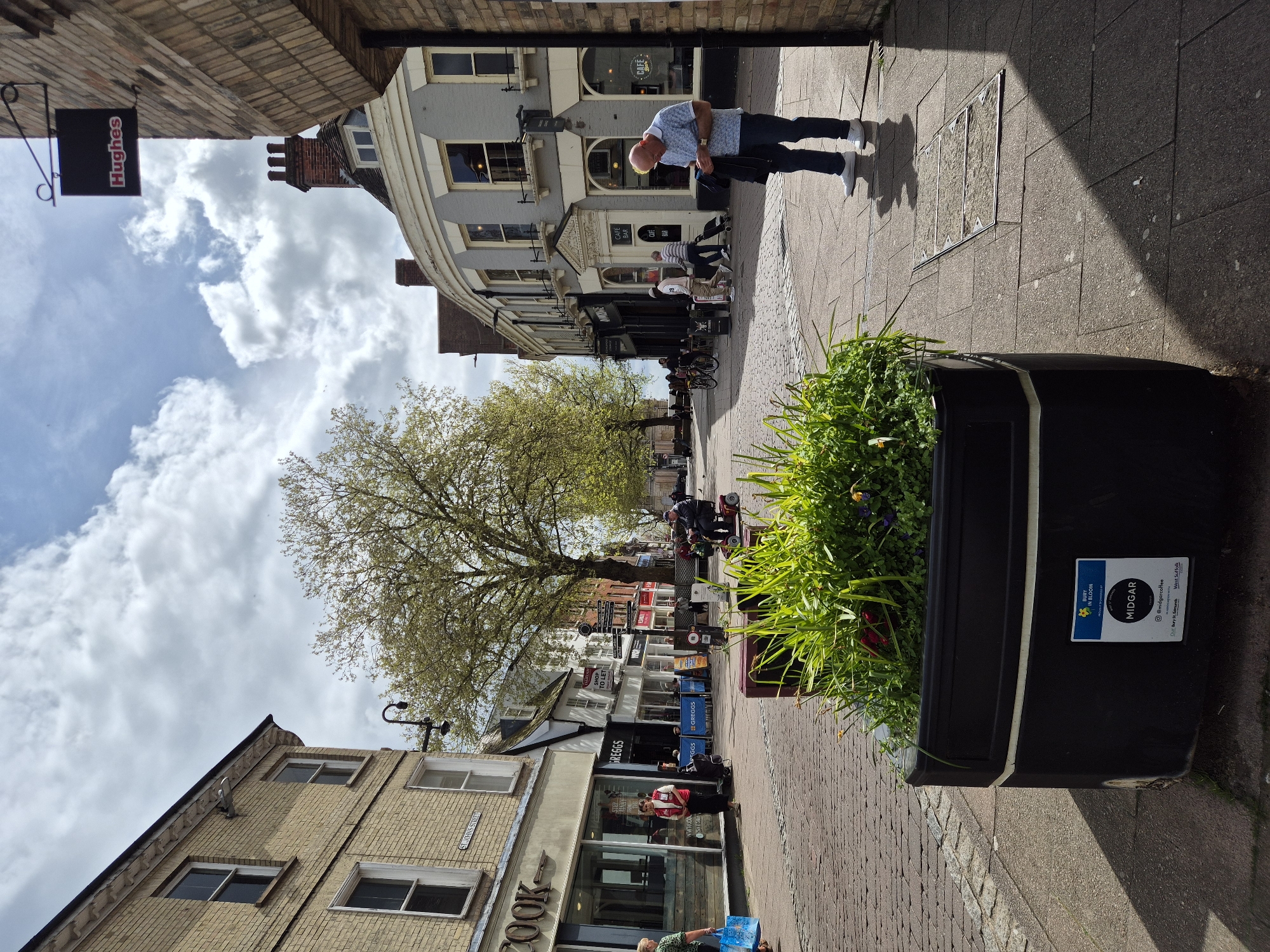 The image depicts a bustling street scene, likely a high street or commercial area, under a partly cloudy sky during daylight hours. The perspective is looking down the street, with buildings lining both sides and a large tree in the middle distance.
Several people are visible, engaged in various activities. A man with a bald head, wearing a light blue patterned shirt and dark trousers, stands prominently on the right sidewalk, looking downwards. Further down the street, individuals are seen walking, standing, and seated at outdoor tables outside cafes and shops. A woman in a red and white jacket stands near a Greggs shop, and another person with a blue shopping bag is near the "COOK" shop. Some people are on mobility scooters in the distance.
The street features traditional multi-story buildings, some with grey facades and bay windows, others with brickwork. Shopfronts are visible along the street. In the immediate foreground on the right, a large black planter filled with lush green foliage and some red flowers is placed on the brick sidewalk. Shadows fall across parts of the pavement, suggesting a bright day.
Visible text includes: "Hughes" on a hanging sign, "COOK" on a shopfront, "GREGGS" and "#GREGGS" on a bakery, and "CAFE BAR" on another establishment. A "SHOP TO LET" sign is also present. On the planter, text reads "BURY IN BLOOM," "MIDGAR," "@midgarcoffee," "Dur Bury St Edmunds," and "West Suffolk." A street sign on a building reads "GARNER STREET."