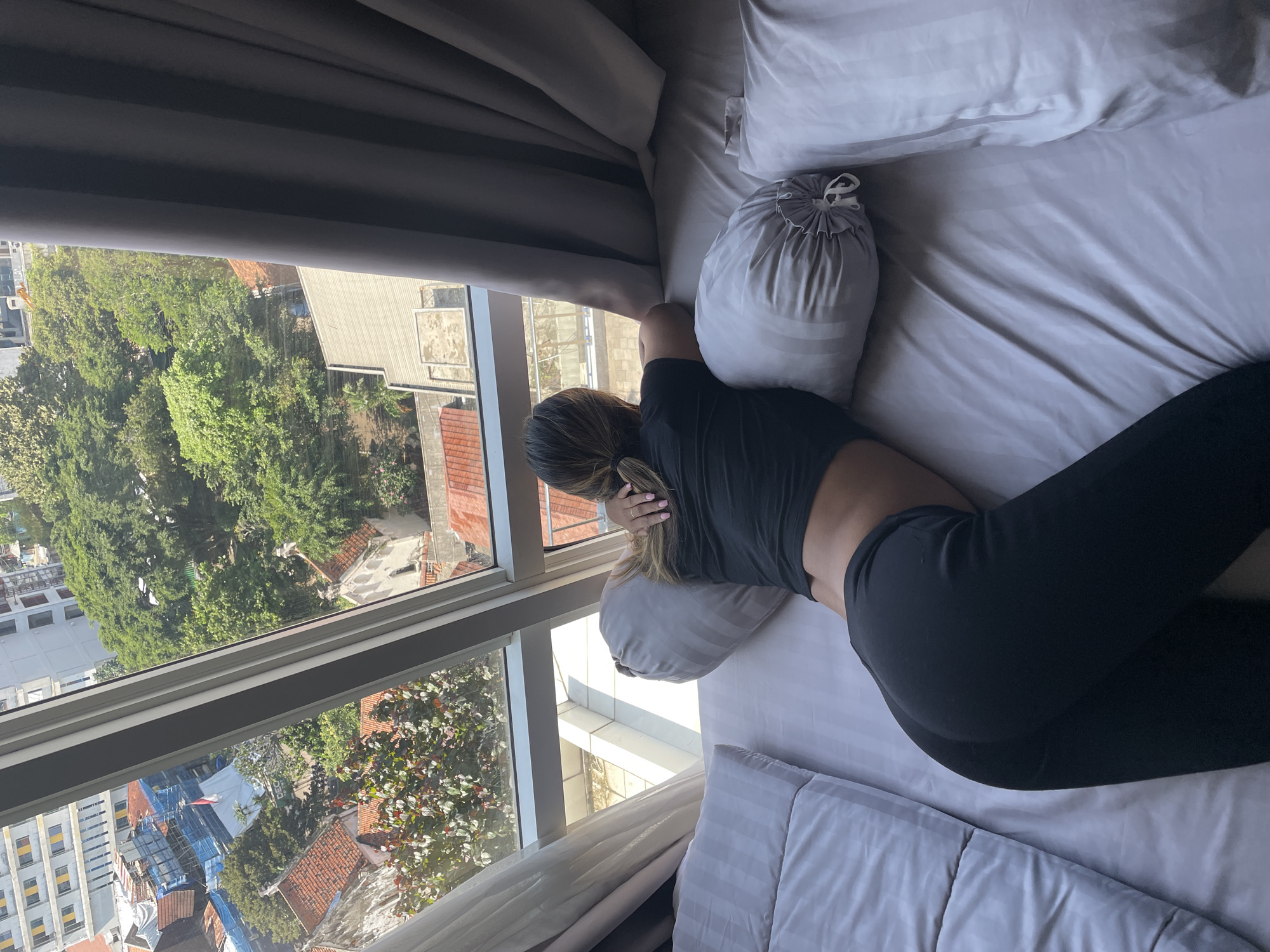 A woman lies on a bed, looking out a window in Central Jakarta, Indonesia. She is wearing a black top and black leggings. The bed has white and gray striped bedding and several pillows. The window looks out onto a cityscape with green trees, buildings, and rooftops. The buildings appear to be a mix of residential and commercial structures. The sky is visible through the window. The angle suggests the photo was taken from a high vantage point. The image is well-lit, indicating daytime.