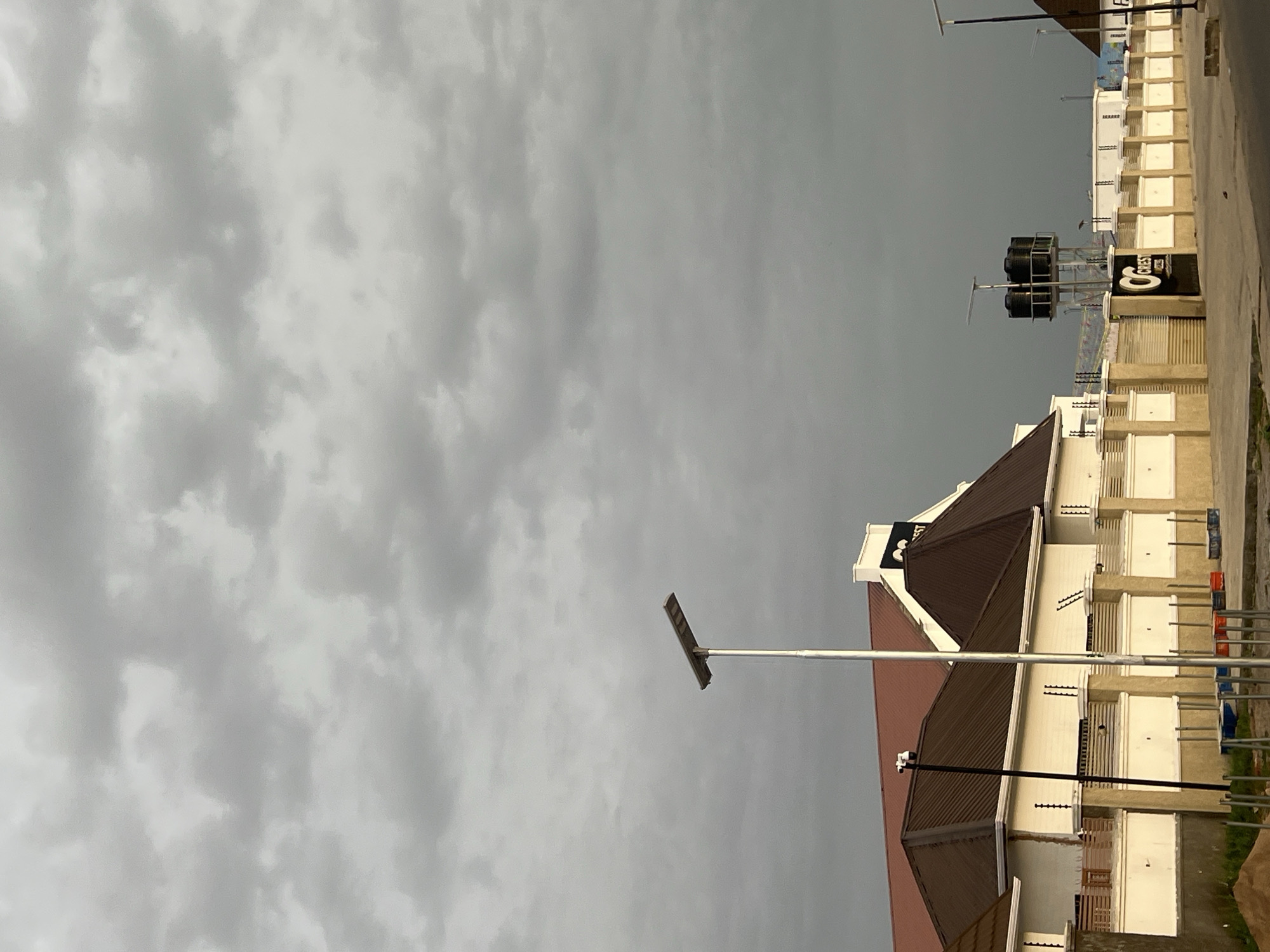 The image shows a street view under a cloudy sky, possibly in Howayi, Nigeria. A two-story building with a brown roof is visible, displaying white walls and numerous windows. Adjacent to the building is another structure with a sign marked with the number "8" in black and white. Several water tanks are elevated behind this building. A long horizontal street light extends out towards the left of the image. The street appears to be paved with concrete. Construction materials and equipment are visible near the base of the building.