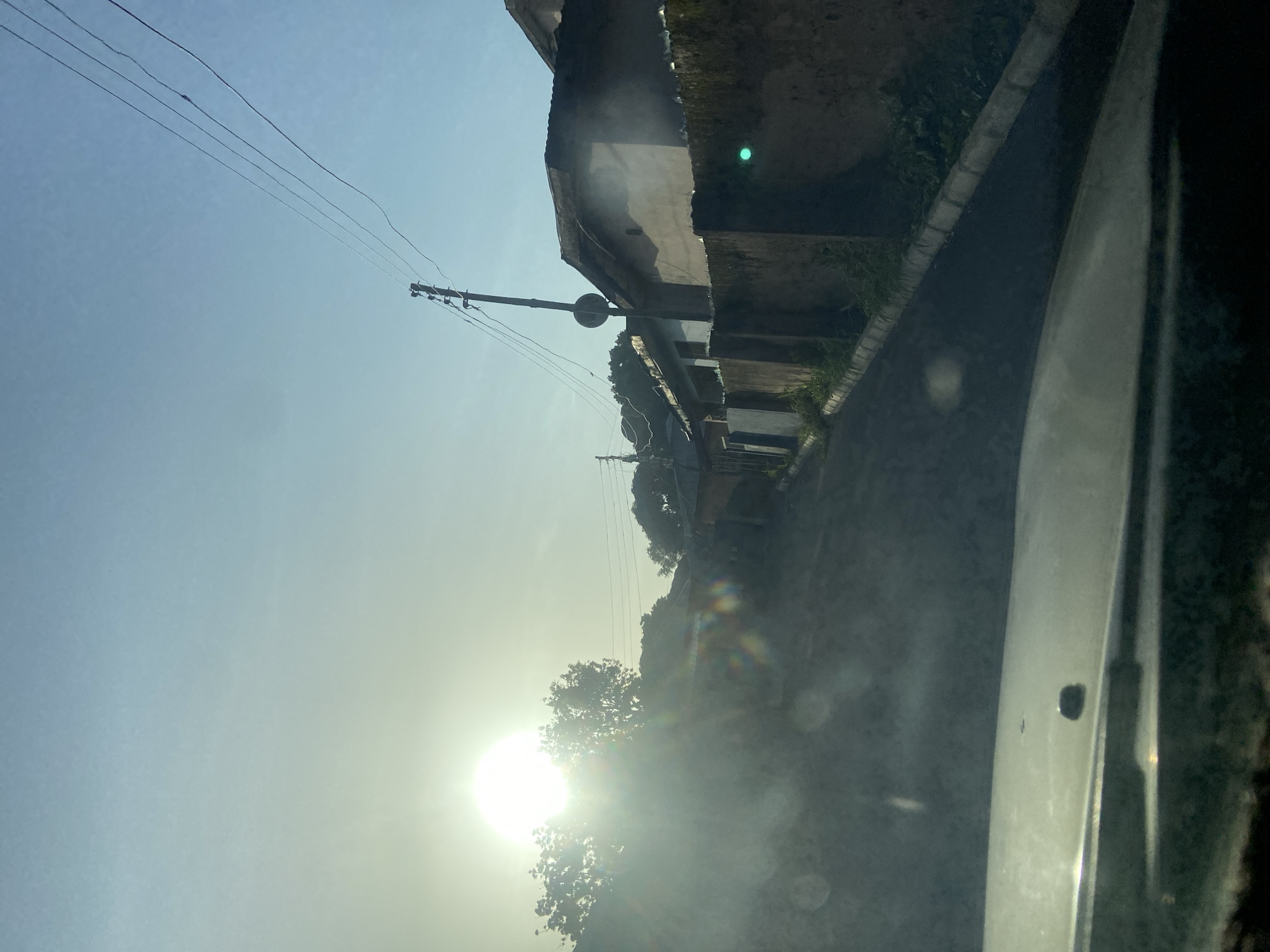 The image depicts a sun-drenched street scene in Jos, Nigeria, likely captured during early morning or late afternoon due to the low angle of the sun and the presence of lens flares. The perspective is from inside a vehicle, looking forward and slightly upwards. A paved road curves gently to the right, bordered by a low, textured wall on the right side, which appears to be constructed from rough-hewn stones or cement blocks. Greenery, possibly weeds or overgrown grass, is visible growing along the base of the wall.
On the right side of the street, several residential buildings with tiled roofs are visible. These structures appear to be single-story or low-rise and exhibit a somewhat weathered appearance. A prominent utility pole with multiple electrical wires running across it stands tall against the bright sky. A circular sign or marker is attached to the pole. Trees, some dense with foliage, are present in the background and to the left of the road, partially obscuring other structures and the terrain behind them.
The sky is a clear gradient of light blue, transitioning to a pale yellow or white near the sun, which is a powerful, bright orb situated in the upper left quadrant of the image. The intense sunlight creates significant lens flares, appearing as circular or oval colored spots scattered across the foreground and middle ground, particularly in the lower left and center areas of the image. The hood of the vehicle from which the photo is taken is visible in the very foreground, taking up the bottom portion of the frame, with a dark, smooth surface. A small dark object, possibly a screw or a debris, is visible on the hood. The overall impression is one of a typical residential street in Jos on a clear, sunny day.