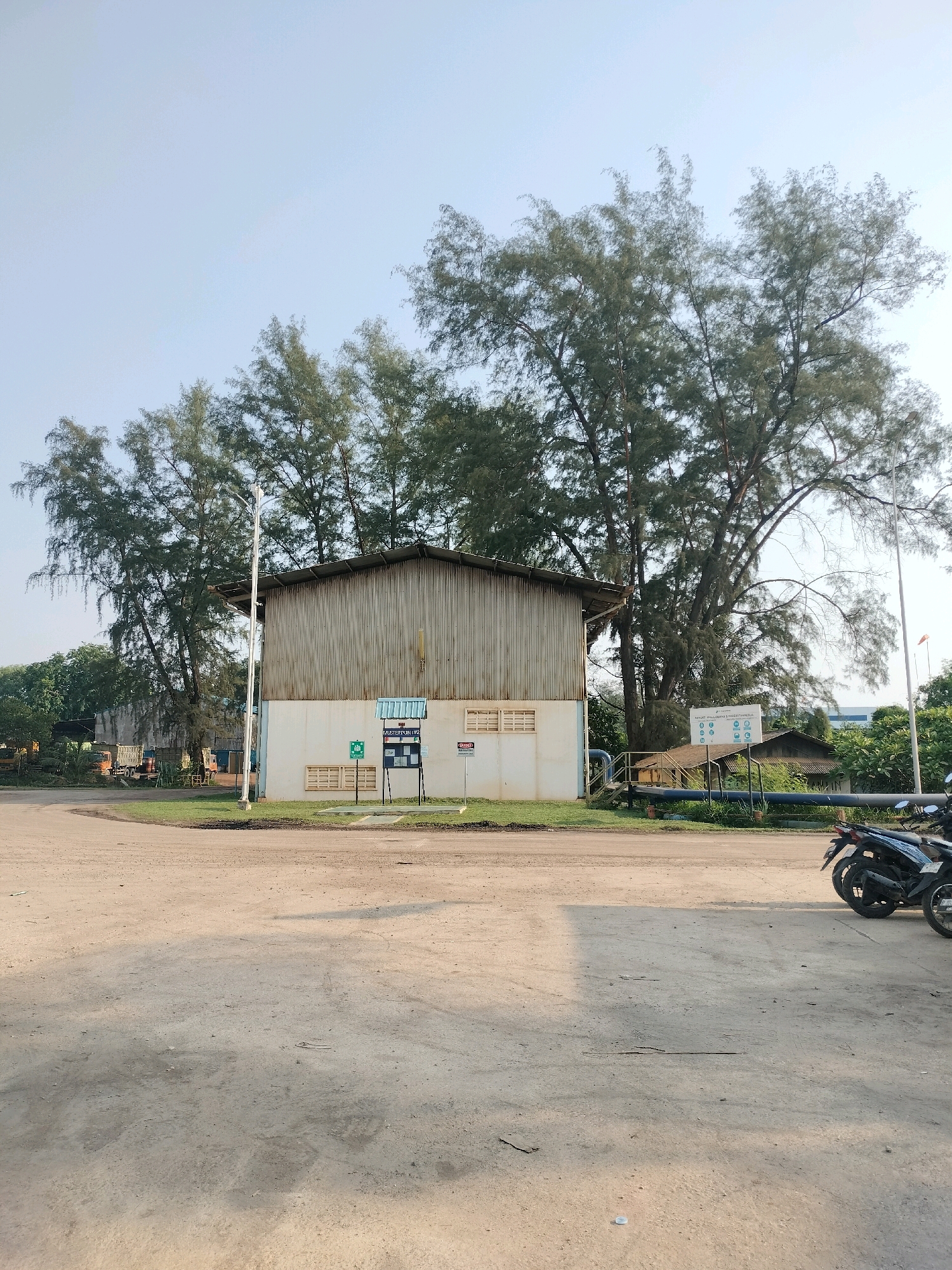A low-angle, outdoor, full shot captures a light brown, unpaved area in the foreground, with a large, light-colored building situated in the midground, partially obscured by tall, mature trees with fine, feathery foliage. The building, with a corrugated metal roof and light-colored walls featuring ventilation grates, appears to be a warehouse or industrial structure. To the left of the building, a white pole with a light fixture stands tall. In front of the building, a blue and white sign is mounted on a post, surrounded by wooden pallets. Further to the left, an orange truck is visible, parked near other vehicles and structures. A network of blue pipes runs horizontally across the midground, connecting to the building and extending towards the right side of the frame. On the far right, two motorcycles are parked. The sky above is a pale, hazy blue. The scene suggests an industrial or logistical area, possibly within a port or industrial complex, common in areas like Batam, Indonesia, known for its manufacturing and trade.