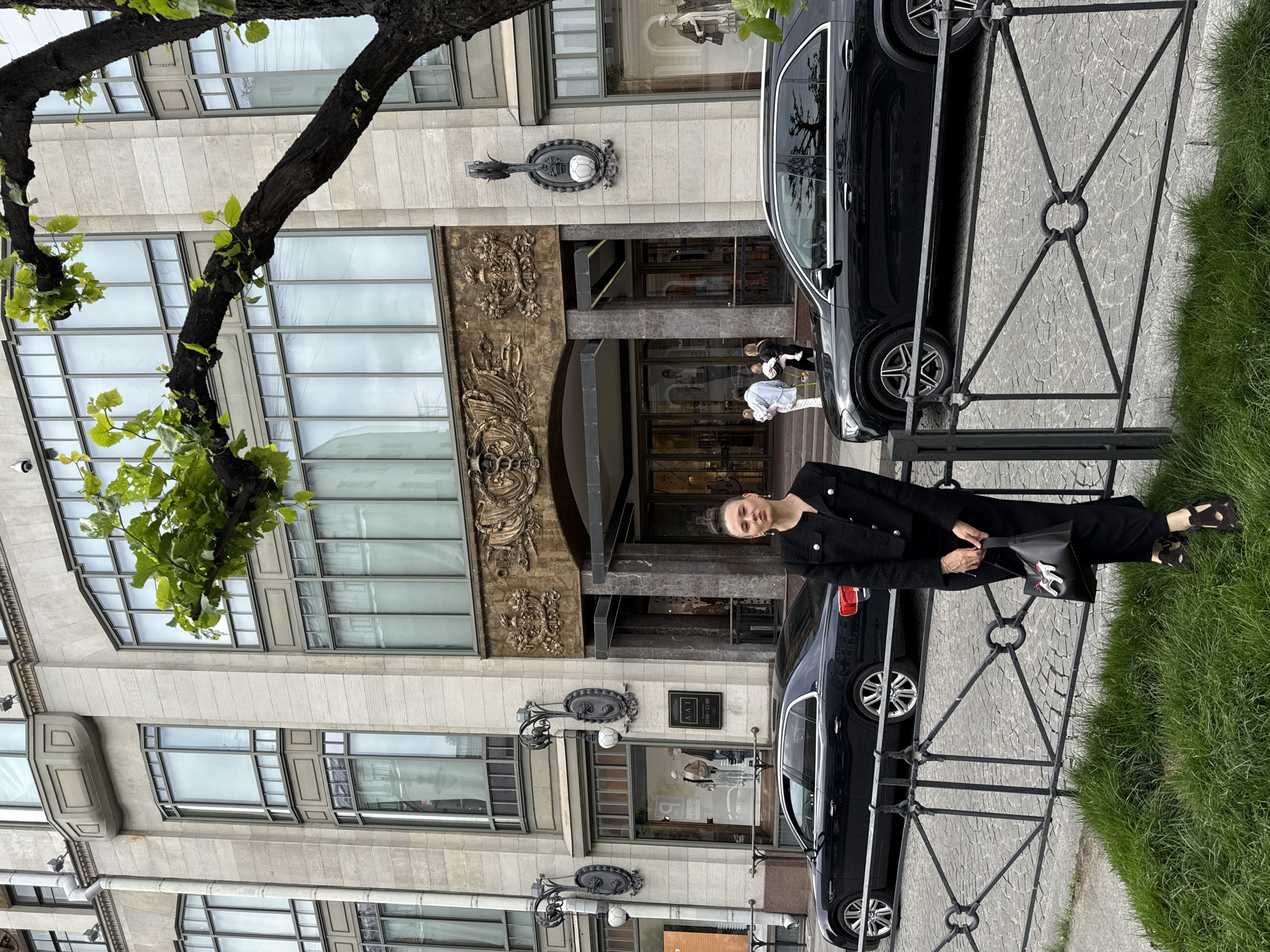 A woman stands in front of a grand building with an ornate entrance in Saint Petersburg, Russia. She is wearing a black textured jacket and pants, with dark sandals and holding a black handbag with a pink charm. Two black cars are parked on the cobblestone street, one a sedan and the other an SUV. In the background, people are seen entering the building, which appears to be a commercial establishment with large display windows. A tree with green leaves partially obscures the upper part of the building and the sky. The woman is positioned behind a decorative metal fence.