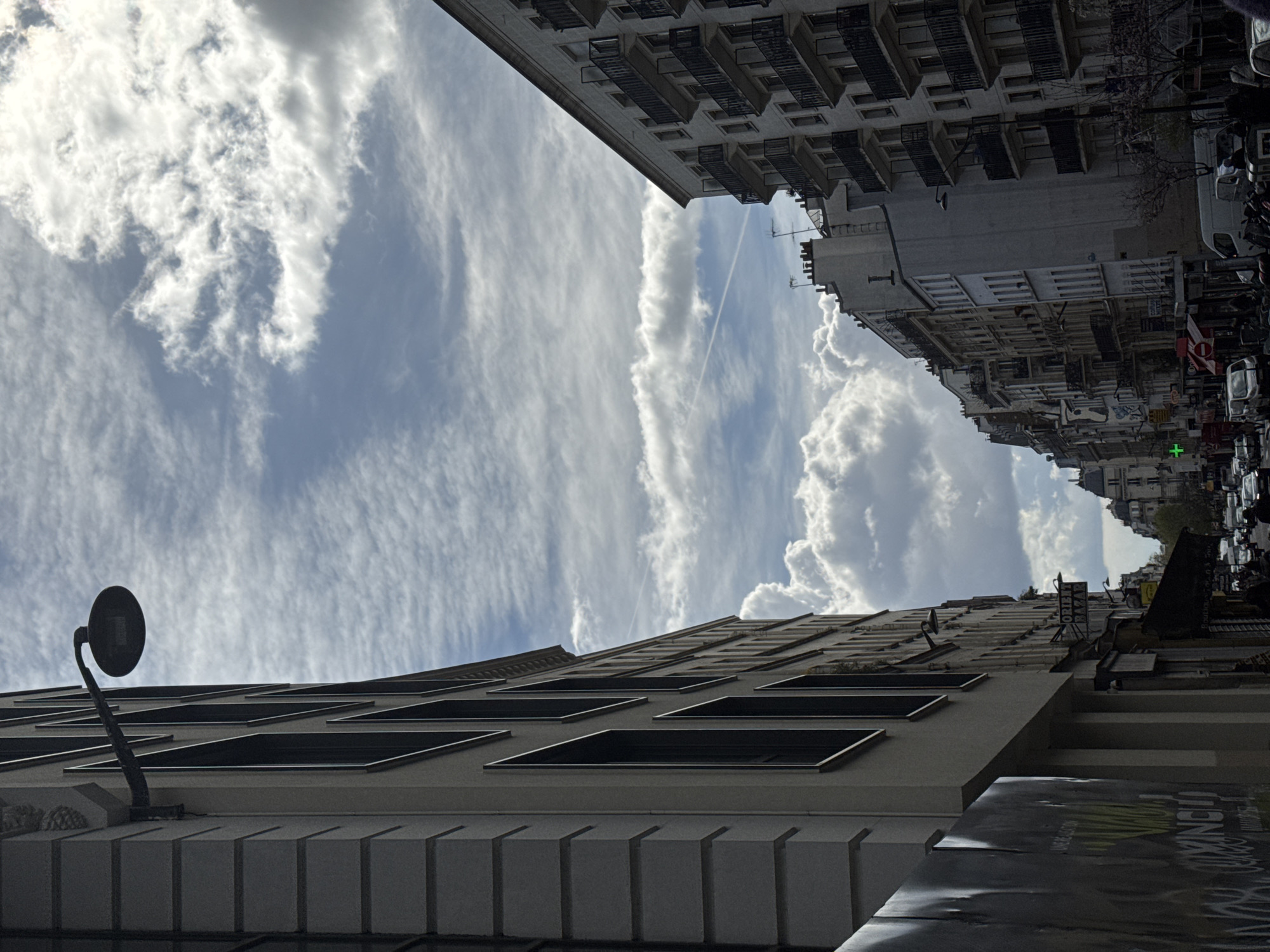 This image captures a street view in Paris, France, looking down a thoroughfare lined with multi-story buildings under a partially cloudy sky. A distinct airplane contrail streaks across the upper-middle sky.
On the left, a tall, light-colored building with a block-textured facade and dark-framed vertical windows occupies the foreground. A dark, circular streetlamp extends from its upper section, casting a silhouette against the sky. The lower portion of this building is obscured by a dark, possibly graffiti-covered construction hoarding or temporary barrier. Further down this side, commercial signs are visible, including one for "ORIENT BAZAR."
On the right, a series of light-colored, multi-story buildings rise, characterized by numerous balconies with dark, decorative iron railings. Some upper-floor windows are open. Rooftops with chimneys are visible along the receding streetline.
The street level is active with vehicles, including cars, vans, and motorcycles/scooters, both parked and moving. Pedestrians are observable on the sidewalks. A prominent green cross sign, indicative of a pharmacy, is mounted on a building further down the street on the left. The overall scene depicts a typical busy urban environment with classic Parisian architecture.