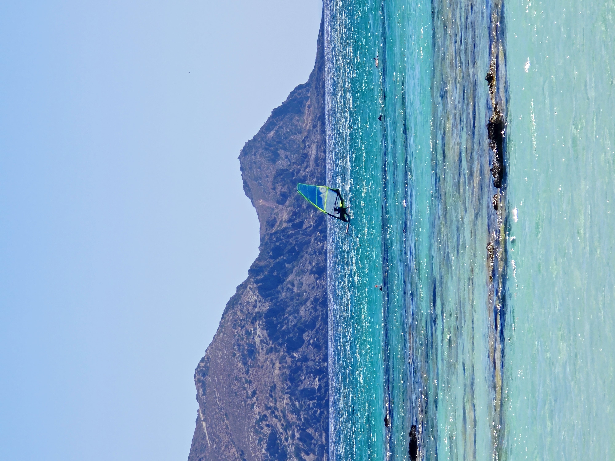 The image depicts a coastal scene in Greece. In the foreground, the clear, turquoise water of the Aegean Sea gently laps against a shallow, rocky seabed. Sunlight glints off the water's surface, highlighting the sandy bottom in places. In the middle ground, a lone windsurfer navigates the vibrant blue waves, their sail a striking triangle of green and blue. Further out, the deep blue of the sea stretches towards a distant, rugged mountain range that dominates the horizon. The mountains are barren and rocky, their slopes casting shadows under the bright, clear sky. The overall impression is one of tranquility and the invigorating beauty of a Greek seascape.