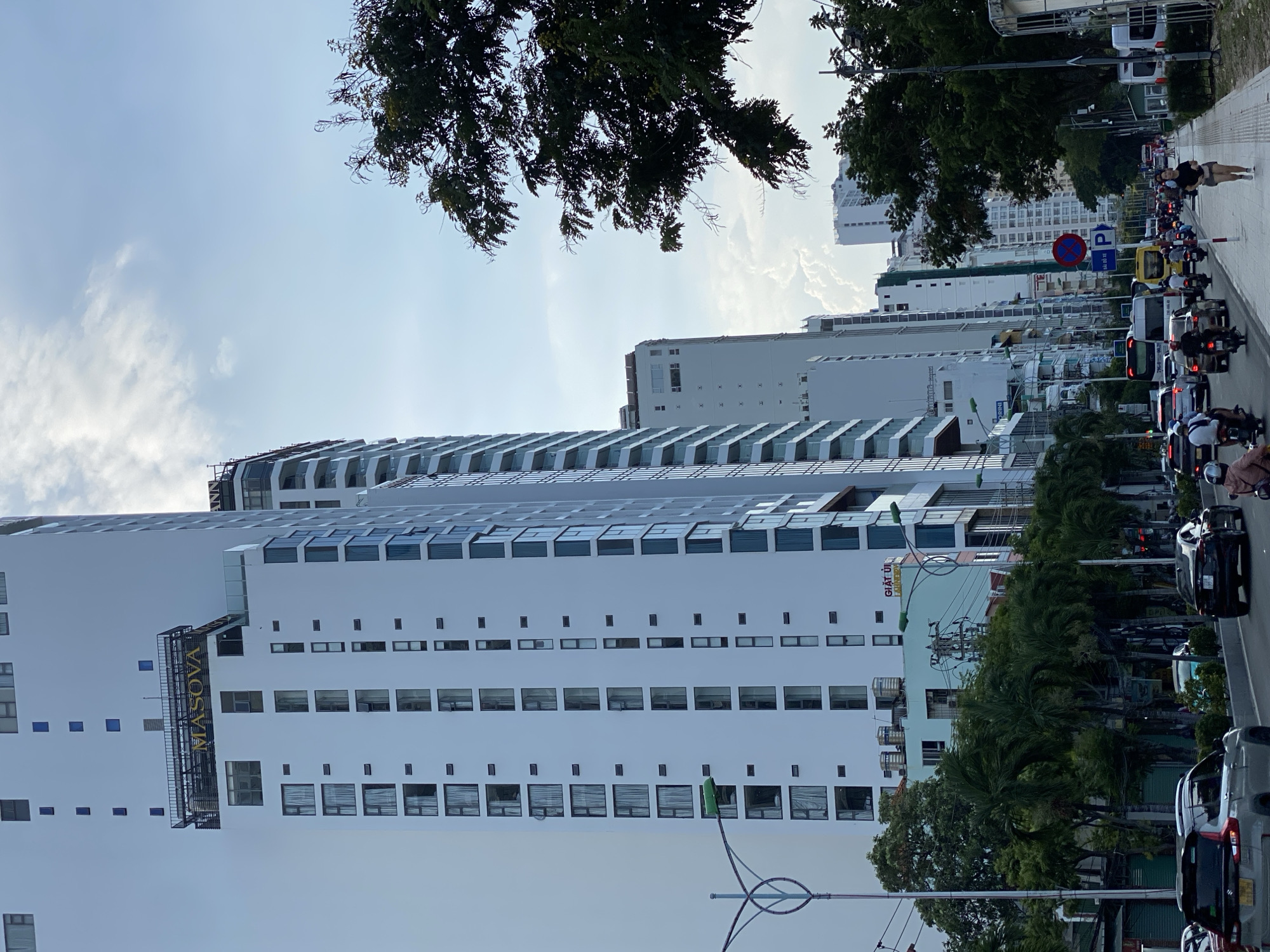 This is a photograph of a street scene in Nha Trang Ward, Vietnam. The image features several tall, modern buildings lining a busy street. One prominent building displays the name "MASOVA" in large gold letters near its rooftop. The buildings are primarily white and glass, with many windows and balconies visible.
On the street, there is moderate traffic, including cars, motorcycles, and what appears to be a bus. Several people are visible, including a woman jogging on the sidewalk, and individuals on motorcycles. Trees, including palm trees, line the street and sidewalk, providing some greenery amidst the urban landscape. The sky is partly cloudy, with a mix of blue sky and white clouds. A street sign with a red circle and a diagonal line over a symbol, along with a blue "P" sign, are visible on the sidewalk. The overall impression is of a vibrant, bustling city environment.