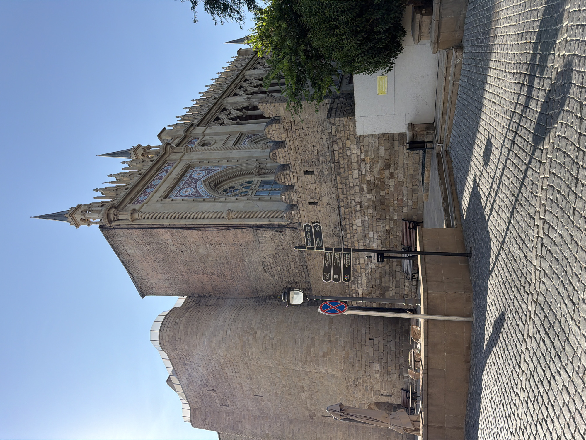 The image depicts a street scene in the Sabail Raion of Azerbaijan, featuring the Palace of the Shirvanshahs. The foreground is a cobblestone street, with a shadow cast from an unseen object stretching across it. To the left, a set of outdoor tables and chairs are arranged under a closed umbrella. A street lamp stands near a sign with multiple directional arrows and text, indicating various locations. Behind these elements, the stone walls of the Palace of the Shirvanshahs dominate the background. The palace has a weathered stone exterior and an ornate facade with intricate carvings and pointed spires. A lush green tree grows to the right of the palace. The sky above is clear and blue.