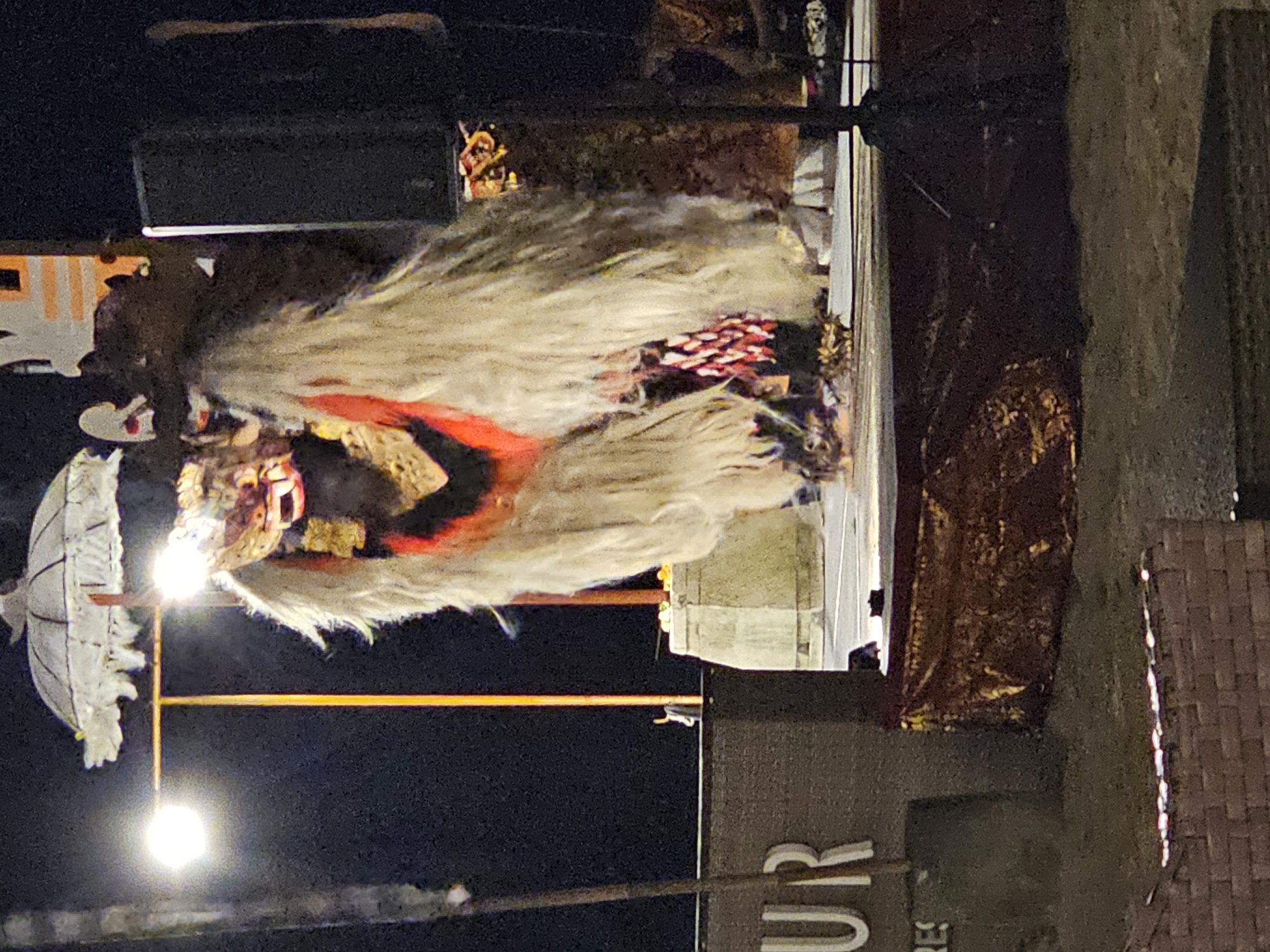The image shows a Barong performance at night in Denpasar, Indonesia. A Barong costume, a mythical lion-like creature, is visible with its ornate mask, white fur, and colorful body. A white umbrella stands behind the Barong. The performance appears to be taking place on a stage or platform with a backdrop. The surrounding environment includes streetlights and building structures.