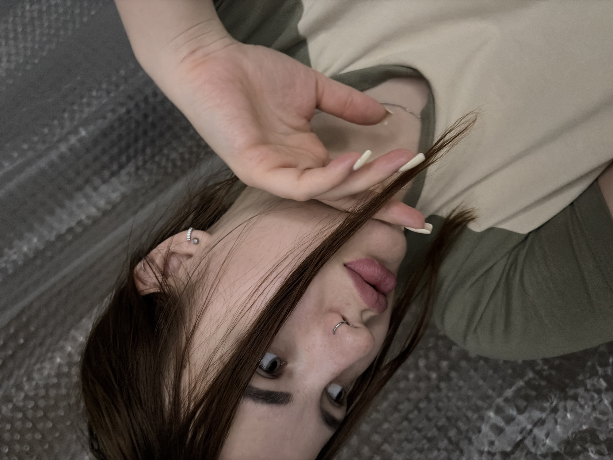 A woman is pictured indoors, lying on a reflective surface. Her head is angled towards the upper left, and she is looking upwards with her eyes open. Her dark brown hair is partially obscuring her face. She has a nose ring in her left nostril and an earring in her left ear. Her hand, with long white nails, is raised and partially covering her face. She is wearing a two-toned shirt, with the top portion appearing to be beige and the bottom portion olive green. There is no discernible information to suggest that this image was taken in Kaluzhskaya, Russia.