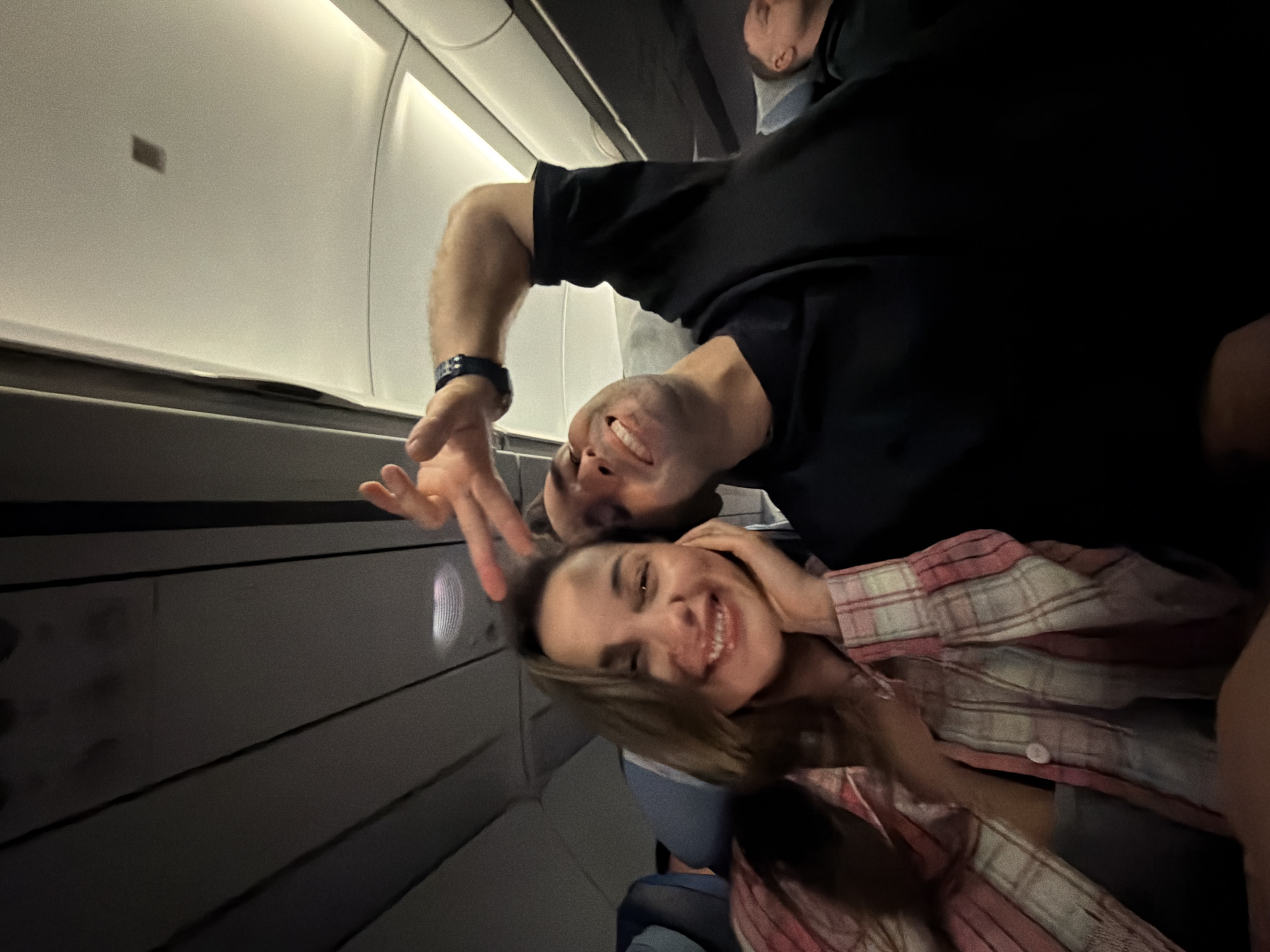 A photo shows two people, likely a couple, posing for a selfie inside an airplane cabin. A man with a black shirt, dark hair, and a wristwatch is partially visible at the top. A woman with long hair and wearing a pink and white plaid shirt is closer to the foreground and smiling. Part of another person is visible above the man. The background reveals parts of the airplane interior, including the windows, wall panels, and overhead storage. The location is likely the Емельяново (аэропорт) in Russia.