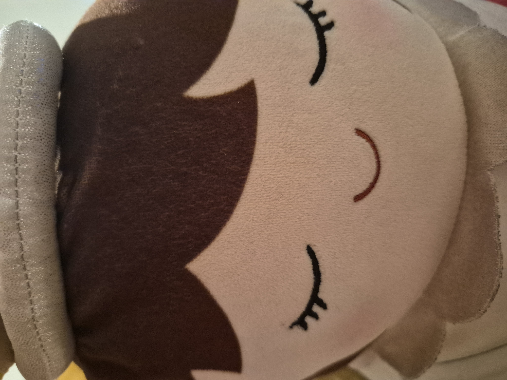 A close-up, frontal view of a stuffed doll's face is presented. The doll has closed eyes, depicted as black curved lines with three short eyelashes on each side. A gentle, curved smile is embroidered in a reddish-brown thread below the eyes. The doll's hair is a dark brown, and it wears a light gray, textured hat or headdress with visible stitching along its top edge. The doll's skin is a light peachy color, and its cheeks have a subtle pink blush. The background is out of focus, showing hints of yellow and white. This image does not contain any information about the location in the city of Sandwell, United Kingdom.