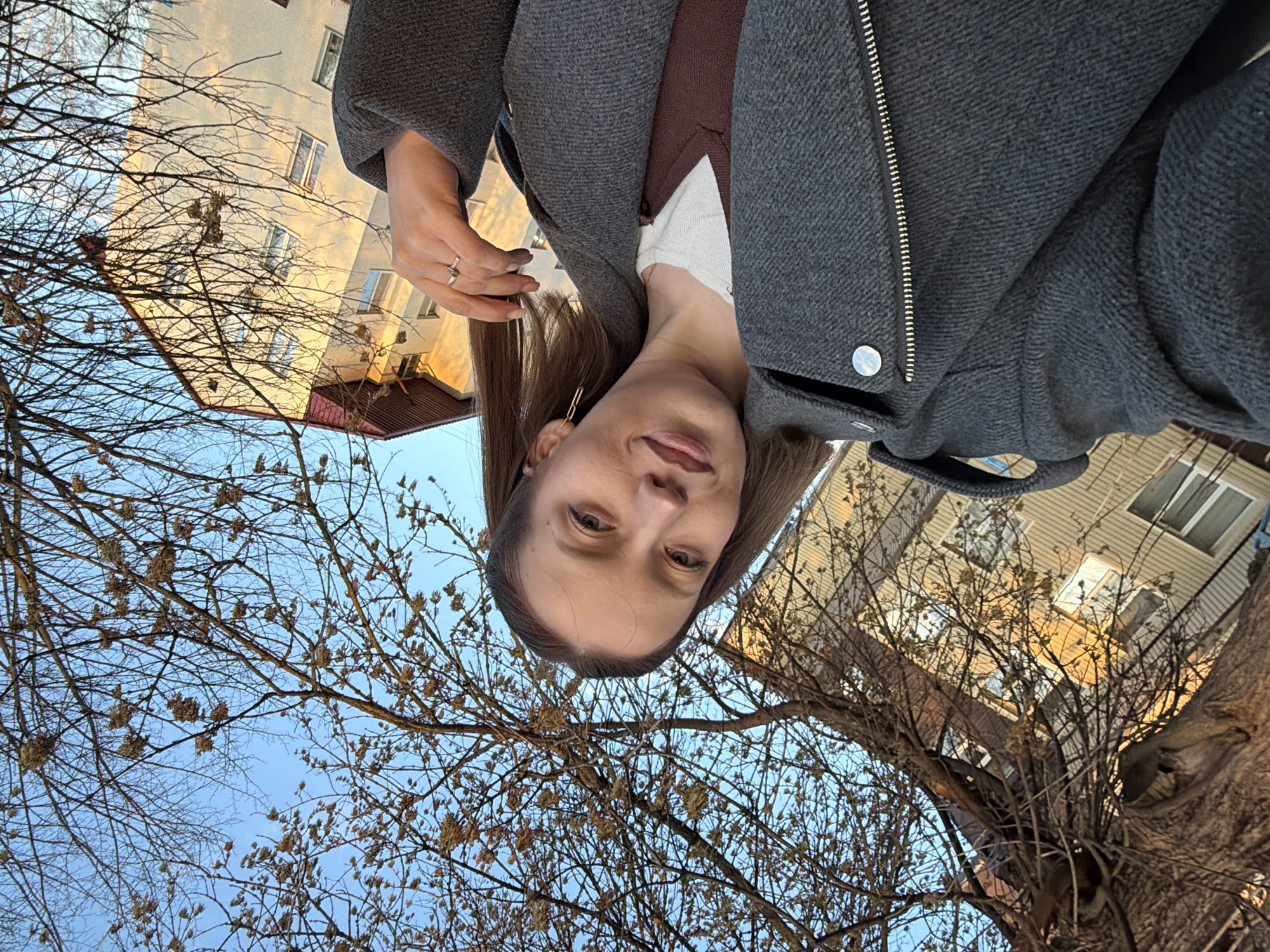 The image is a low-angle selfie of a woman in what appears to be an urban setting, likely in Reshetnikovo, Russia, given the prompt. The woman is wearing a gray coat over a brown top with a white undershirt. A metal zipper runs down the front of the coat, and a ring is visible on one of her fingers.
Above and around her are bare branches of a tree against a backdrop of a pale blue sky. Buildings are visible in the background, some with windows. The overall impression is that the photograph was taken outdoors in an urban environment, likely during the fall or winter season when trees have lost their leaves.