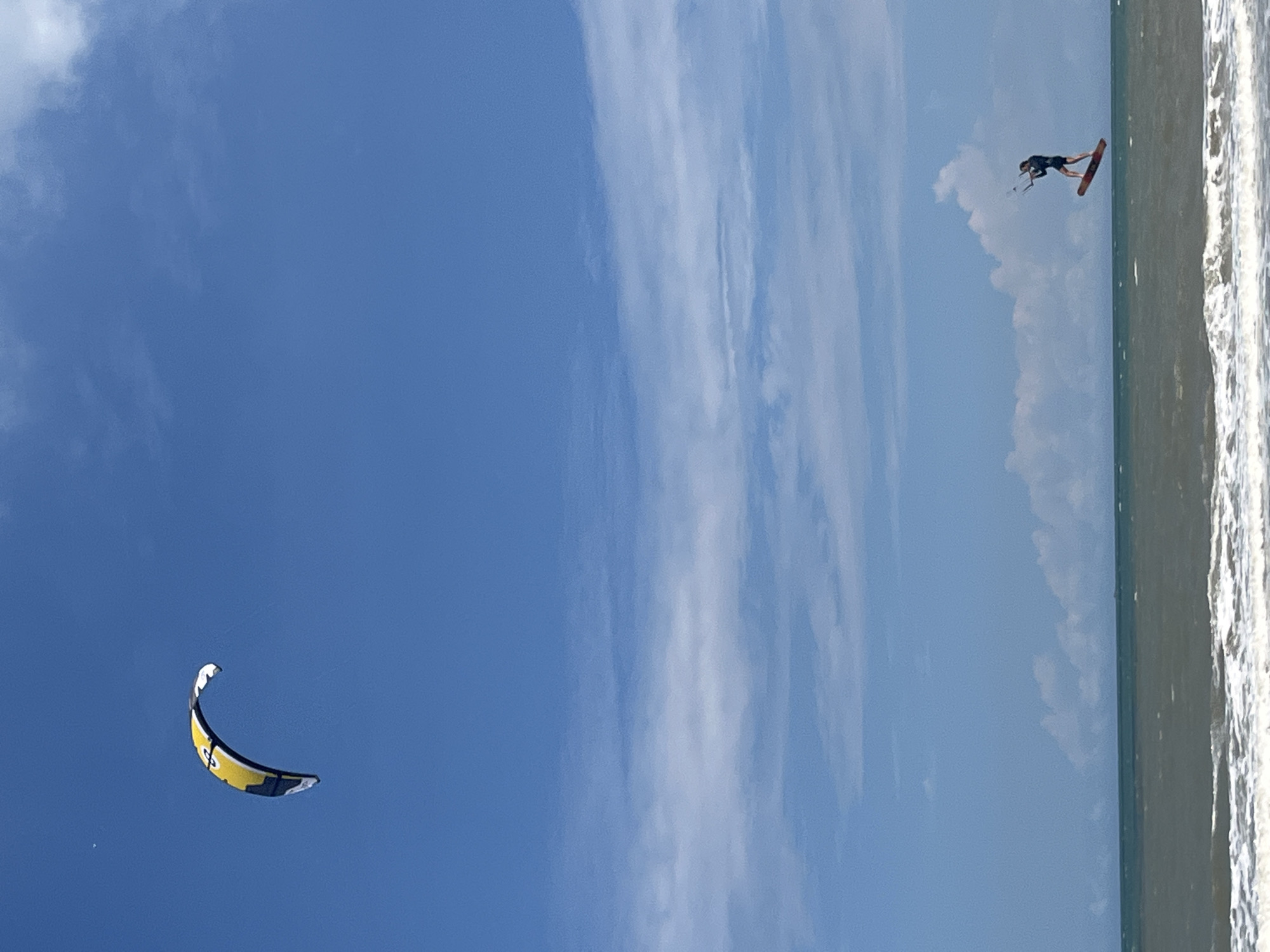 A kitesurfer is captured mid-air over a body of water. The kitesurfer, wearing dark athletic wear, is on a board and appears to be performing a trick, with their body extended. A large, yellow and black kite, curved like a crescent moon, is visible in the sky to the left. The sky is a clear blue with scattered white clouds. To the right of the image, the ocean is visible, with waves crashing against a sandy shore. The water is a greenish-blue color. The scene suggests a sunny day, likely daytime, with good weather conditions for kitesurfing. The location is Phu Thuy Ward, Vietnam. There is no visible text in the image.