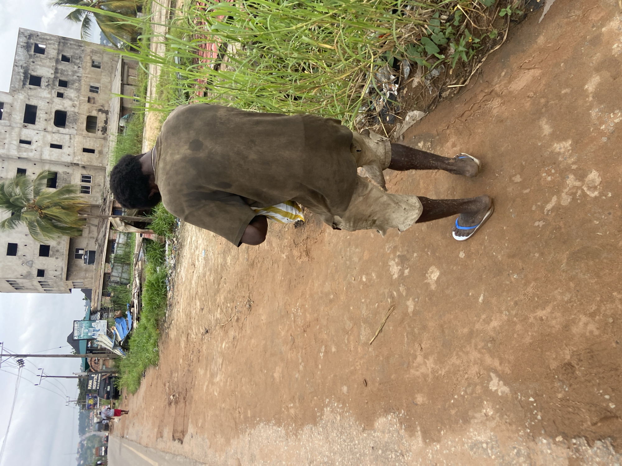 In Nkpor, Nigeria, a man walks along a dirt road. He wears a faded, dark t-shirt and light-colored shorts, with a white and yellow plastic bag in his right hand. He has blue and white sandals on his feet. The road is unpaved and dusty, with patches of wetness indicating recent rain. To the right of the road, tall, green grass grows along a littered embankment, with discarded items visible. In the background, a partially constructed, multi-story building stands, appearing weathered and abandoned, with palm trees nearby. Further down the road, another person in red walks away from the camera, and several vehicles, including a black SUV, are present. Power lines and poles are visible, typical of urban infrastructure. The sky is overcast, suggesting a cloudy or possibly rainy day.