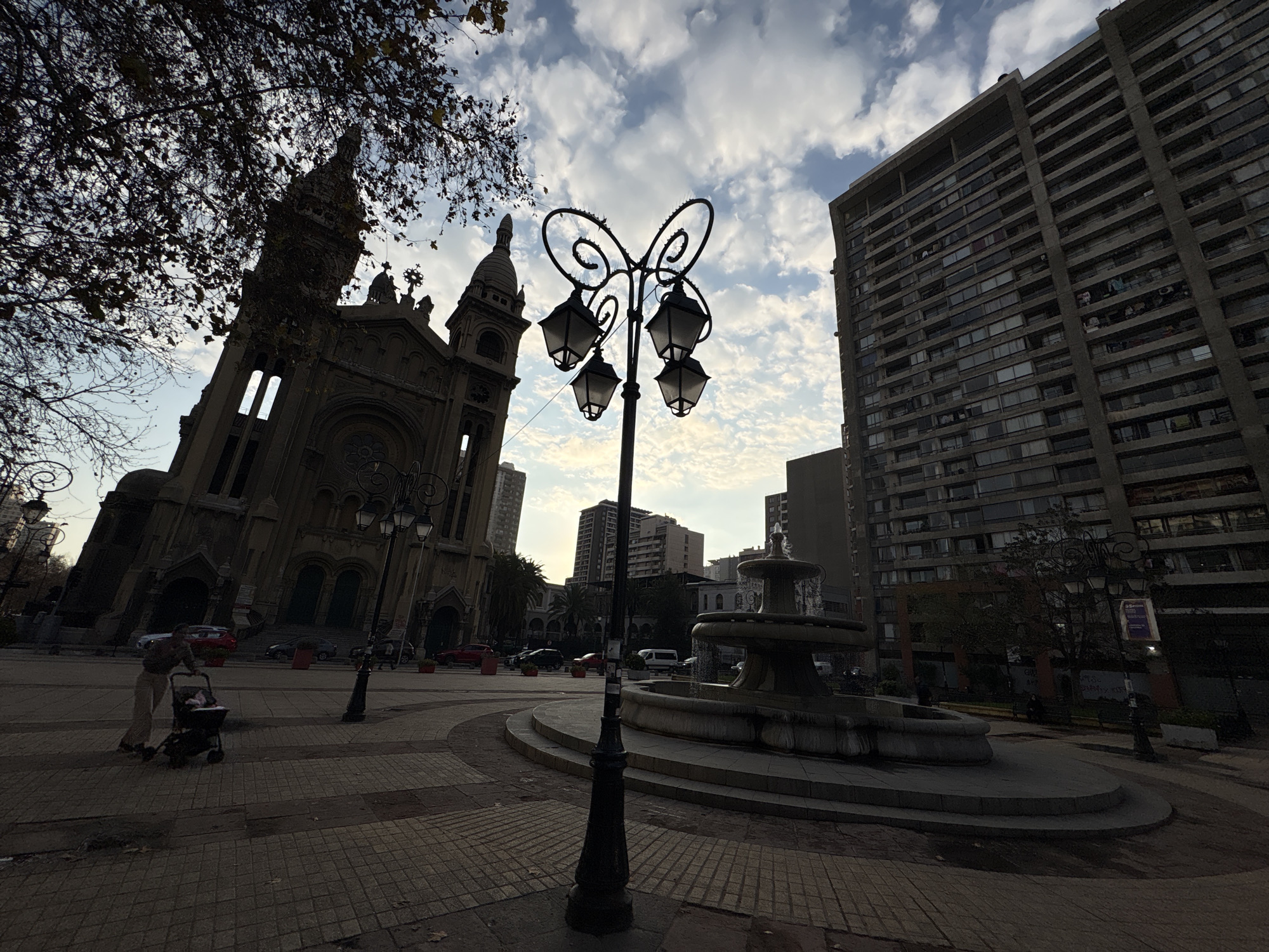 The image depicts an urban square in Santiago, Chile, featuring a mix of historic and modern architecture. In the foreground, a vintage street lamp with four lamps and a decorative butterfly-like design stands prominently. To the left, a large, ornate church, likely Basilica del Salvador given the architectural style and Santiago setting, is visible with its spires and arched windows. In the background, a tall, brutalist-style apartment building dominates the skyline, a common sight in Santiago. A circular fountain with multiple tiers is situated in the mid-ground, with water flowing. A person is pushing a stroller on the paved walkway in front of the church. The ground is paved with interlocking bricks, and trees with bare branches, suggesting autumn or winter, are present. The sky is partly cloudy, with the sun setting or rising, casting long shadows. The overall scene suggests a public space in a bustling city, with elements of historical significance and contemporary urban development.