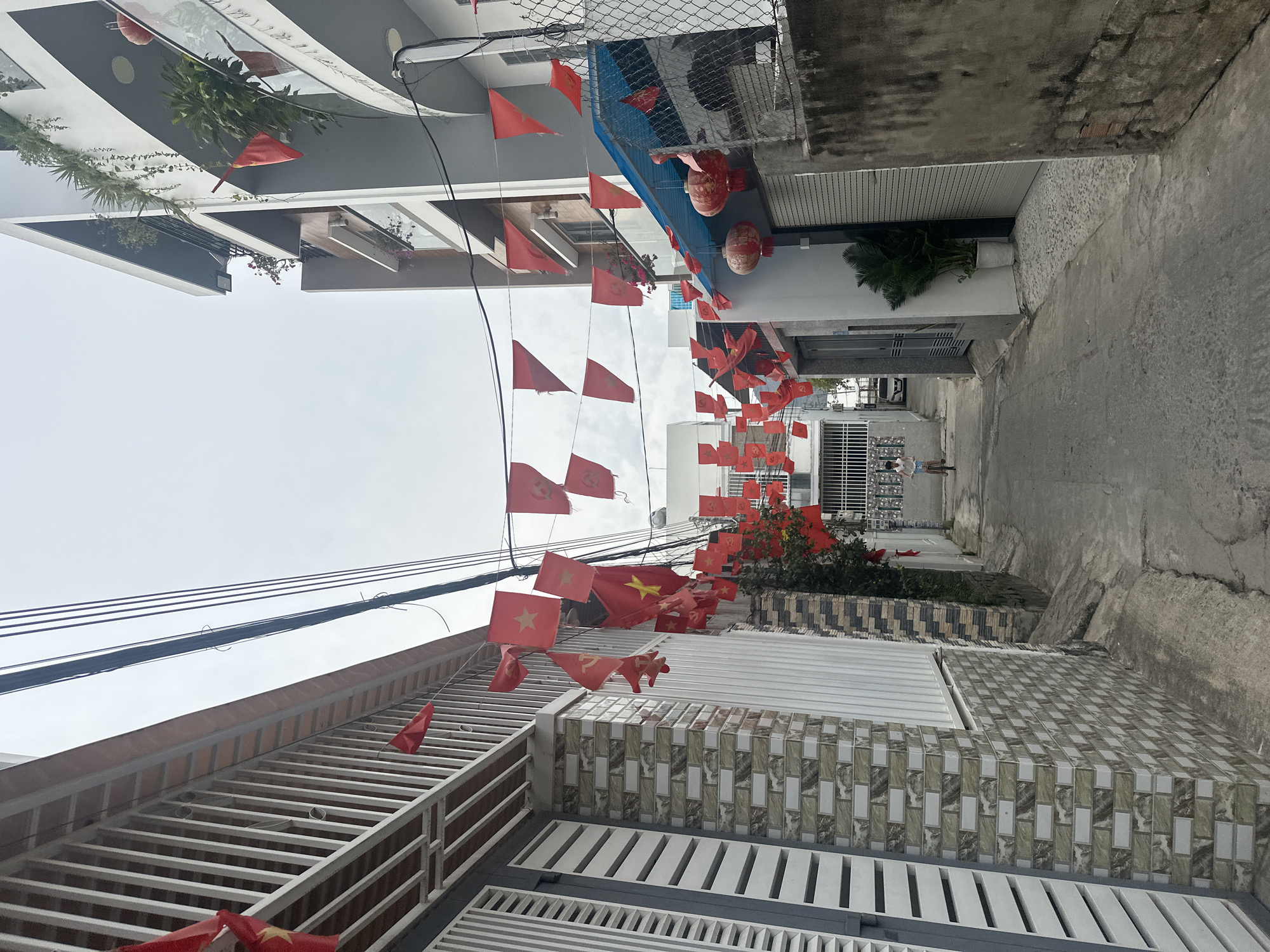 The image captures a narrow alleyway in Bac Nha Trang Ward, Vietnam. The alley is lined with buildings of varying heights and architectural styles. Red Vietnamese flags are strung across the alleyway, creating a festive atmosphere. Red lanterns are visible as well. Two young people can be seen walking away from the camera at the far end of the alley. Electrical wires crisscross the sky above the alley. The buildings appear to be constructed with a mix of materials, including concrete, metal, and patterned tiles.