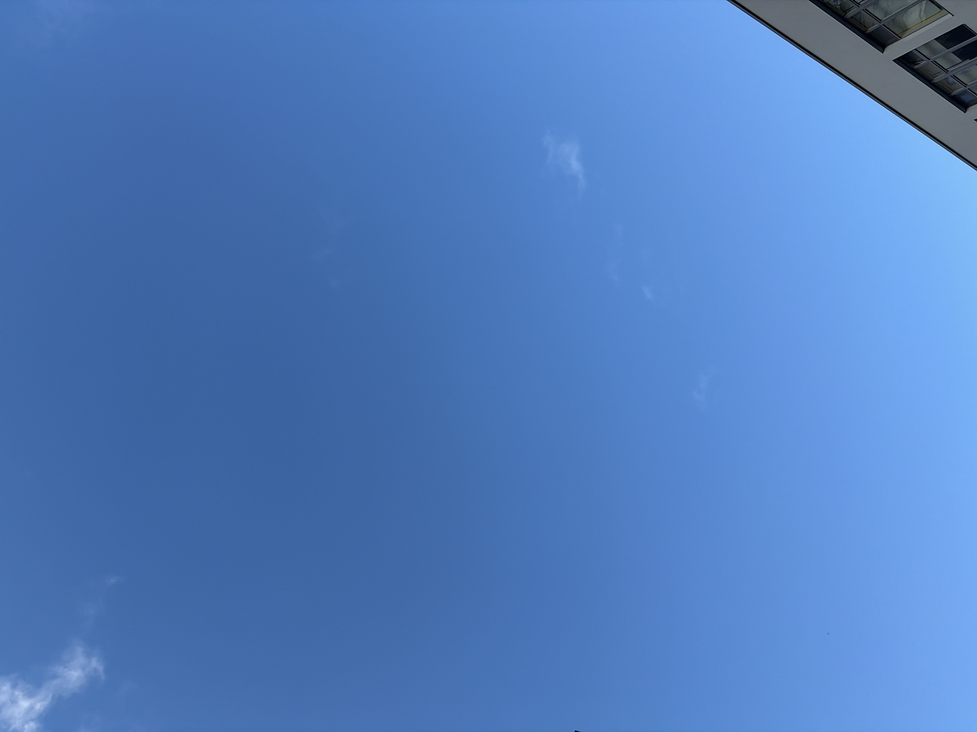 The image is a low-angle shot of a clear, blue sky with a few small, wispy clouds. In the upper right corner, a section of a building is visible, featuring a white facade and gridded windows.