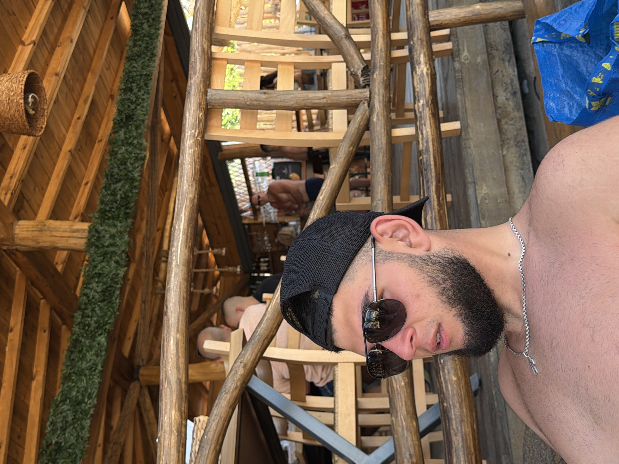The image shows a young man in the foreground, seated, wearing a black baseball cap and sunglasses. He has a beard and is wearing a silver chain around his neck. He is shirtless. Behind him, there are other people seated at tables and standing in what appears to be an outdoor dining or bar area with wooden structures and a thatched roof. The setting is likely a beachside establishment in Primorsko, Bulgaria, given the context. A blue IKEA bag is visible on a wooden bench to the right of the man. The environment suggests a casual, summer atmosphere.